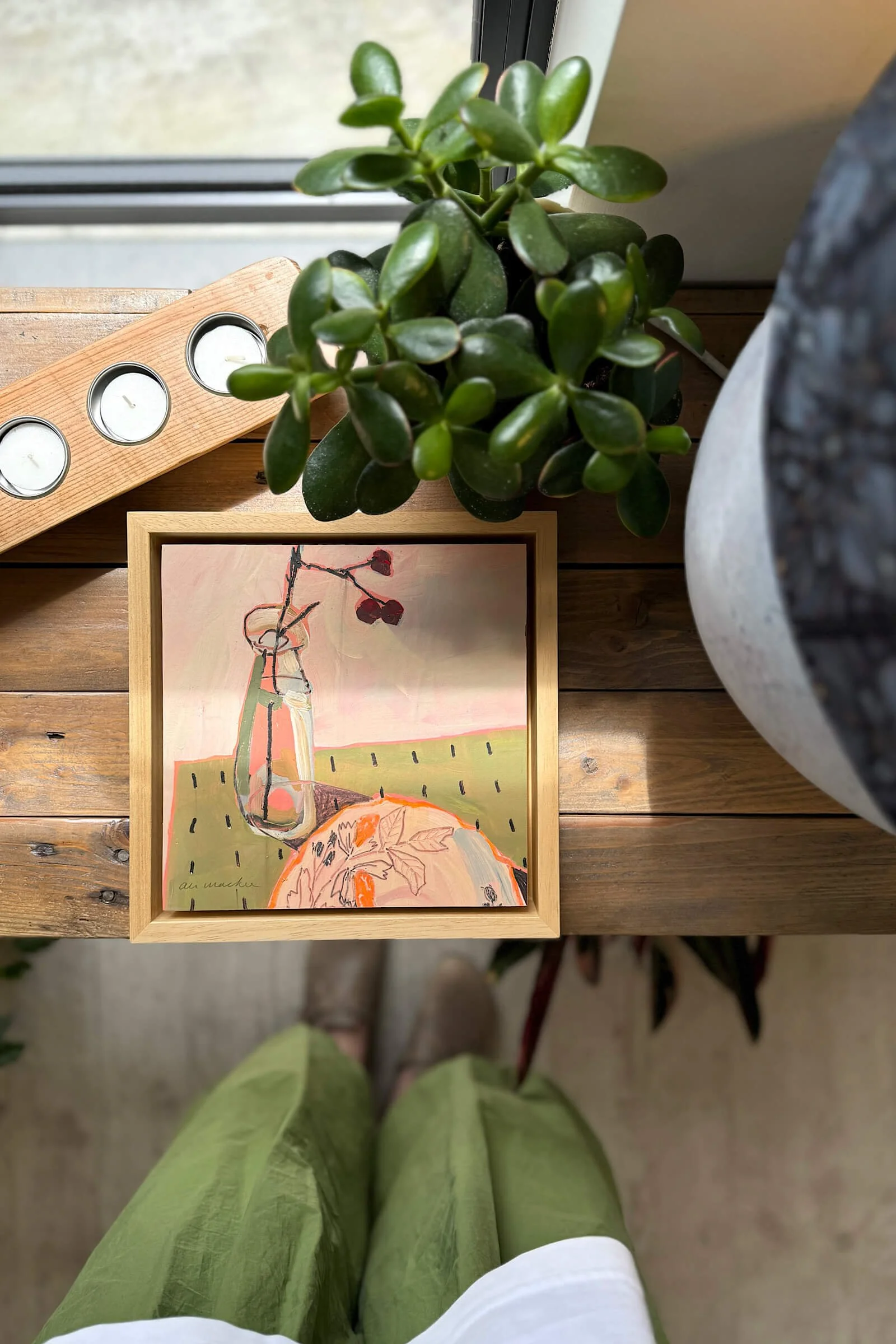 A wooden table holding a framed art piece, a potted plant, and a wooden candle holder with four tea light candles. The photo is taken from a top-down perspective, showing the person's green pants and shoes from above.