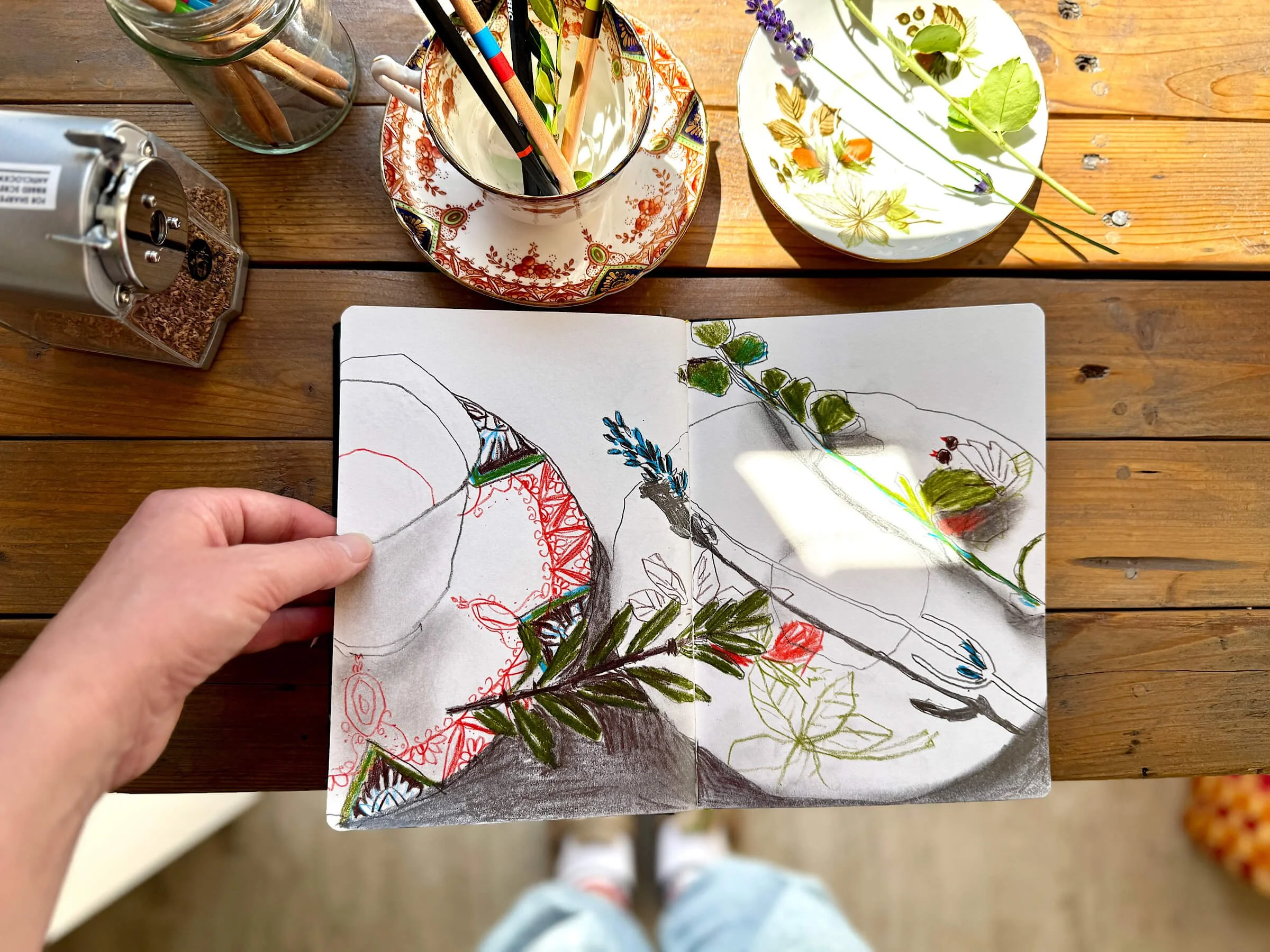 Top-down view of an open sketchbook on a wooden table, featuring colorful pencil drawings of teacups, saucers, and botanical stems, surrounded by real teacups, pencils, and fresh lavender.