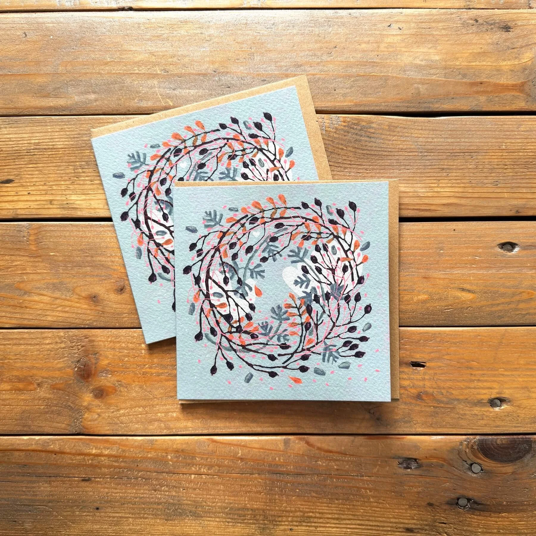 Pair of blue and coral festive greeting cards featuring an original hand-painted botanical wreath artwork, shown on rustic kraft envelopes.