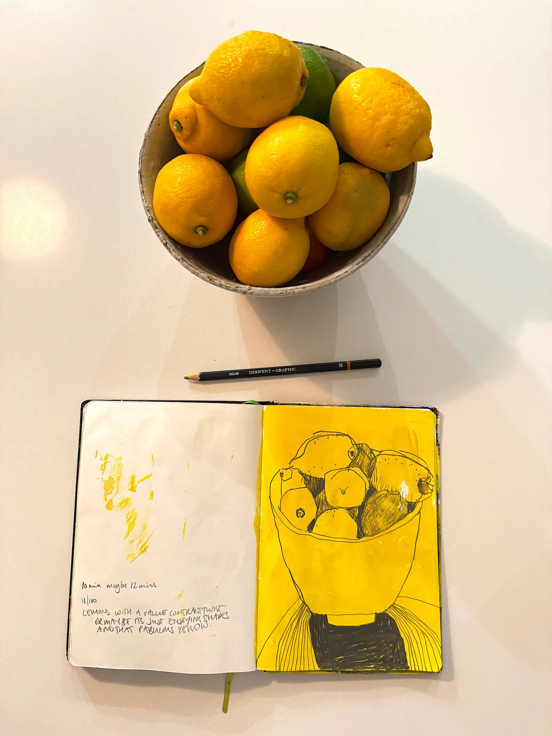 A top-down view of an open artist sketchbook on a white table displaying a vibrant yellow ink line drawing of a bowl of lemons, placed directly underneath a real ceramic bowl filled with fresh yellow lemons and a graphite pencil.
