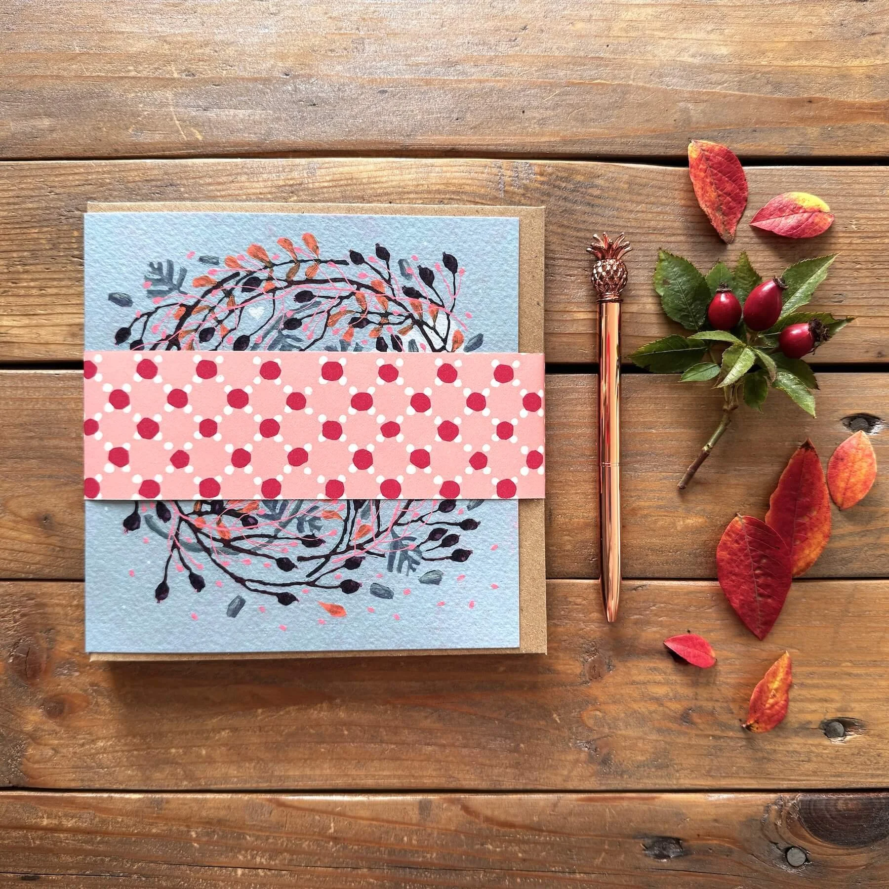 Pack of 8 Wreaths festive greeting cards wrapped in a polka-dot paper belly band with kraft envelopes on a wooden background with autumn leaves and rose gold pen.