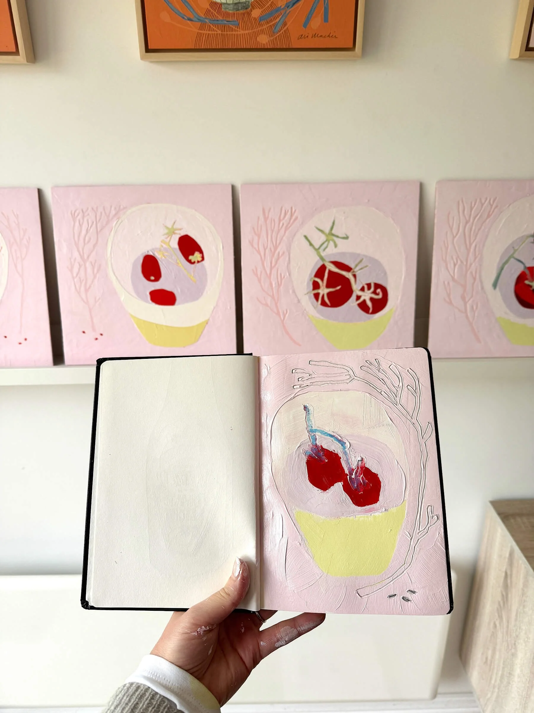 An artist's hand holds up an open sketchbook showing a textured pink and red tomato drawing, positioned directly in front of a row of square contemporary still life paintings on board featuring red vine tomatoes on soft pink backgrounds.