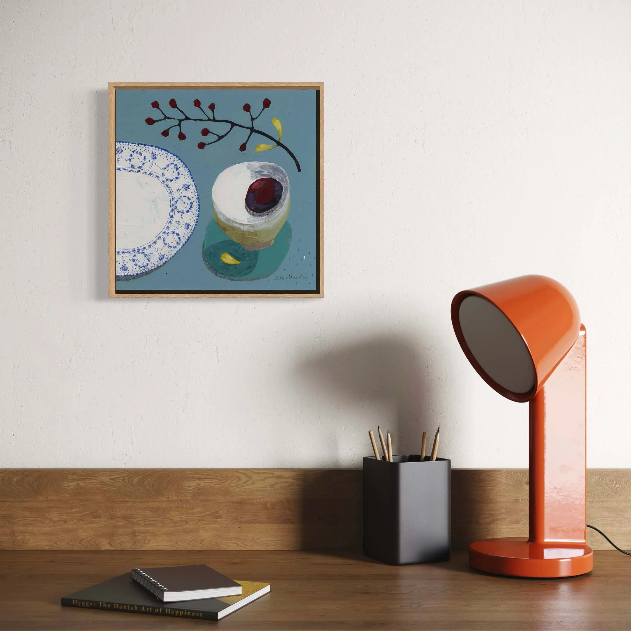 Square framed botanical still life painting by Ali Mackie Art hanging above a desk with an orange desk lamp.