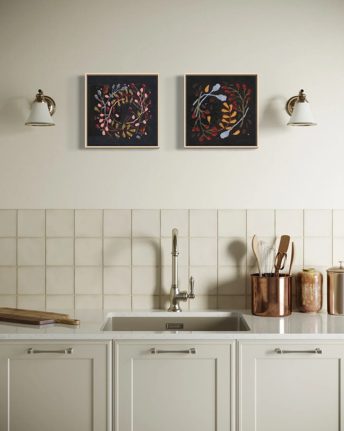 A pair of dark framed botanical wreath paintings hanging side by side on a neutral wall above a kitchen sink and tiled backsplash.