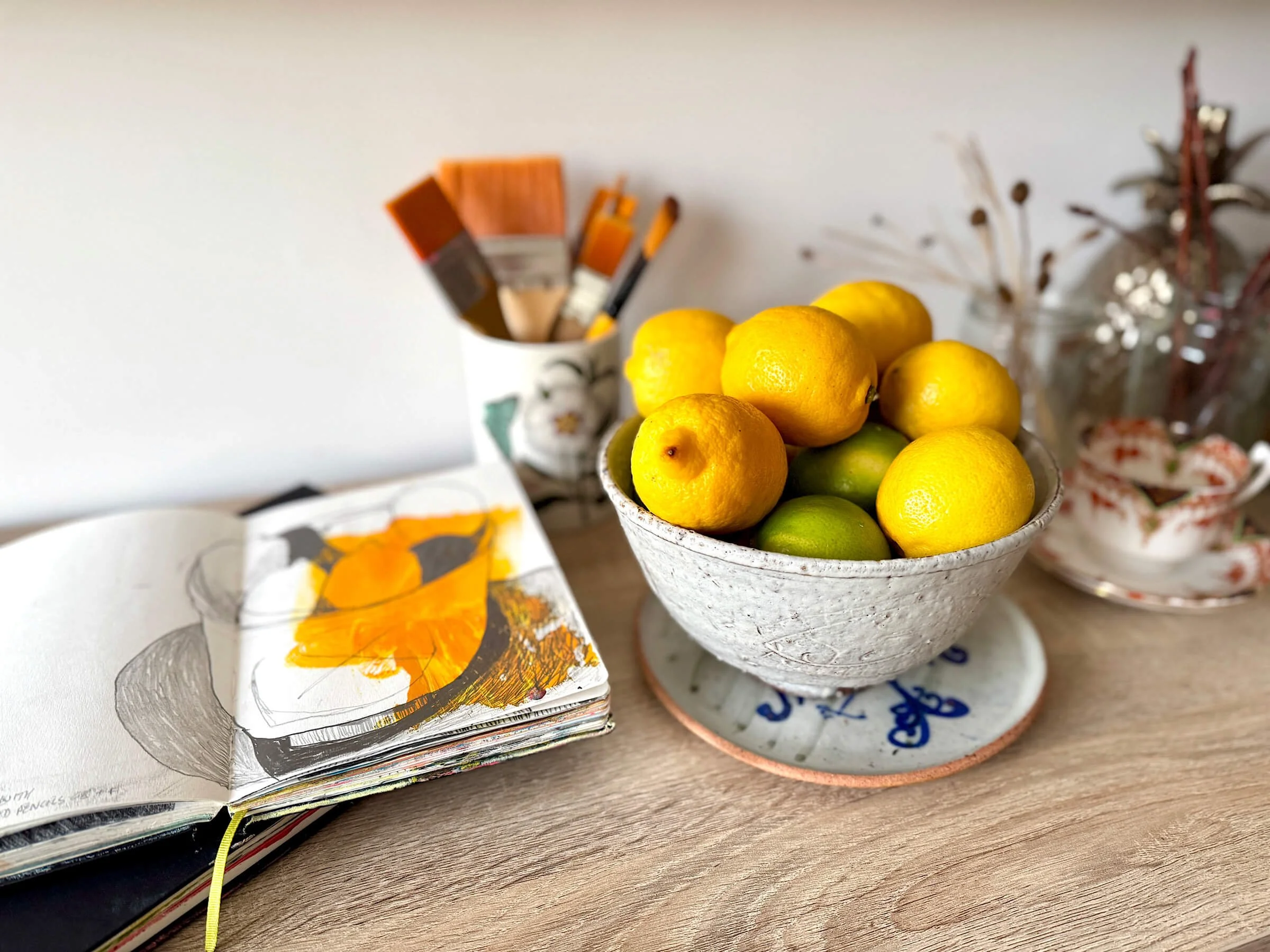 A close-up view of Ali Mackie Art’s studio sideboard featuring a white ceramic bowl filled with bright yellow lemons alongside vintage floral teacups and patterned porcelain saucers.