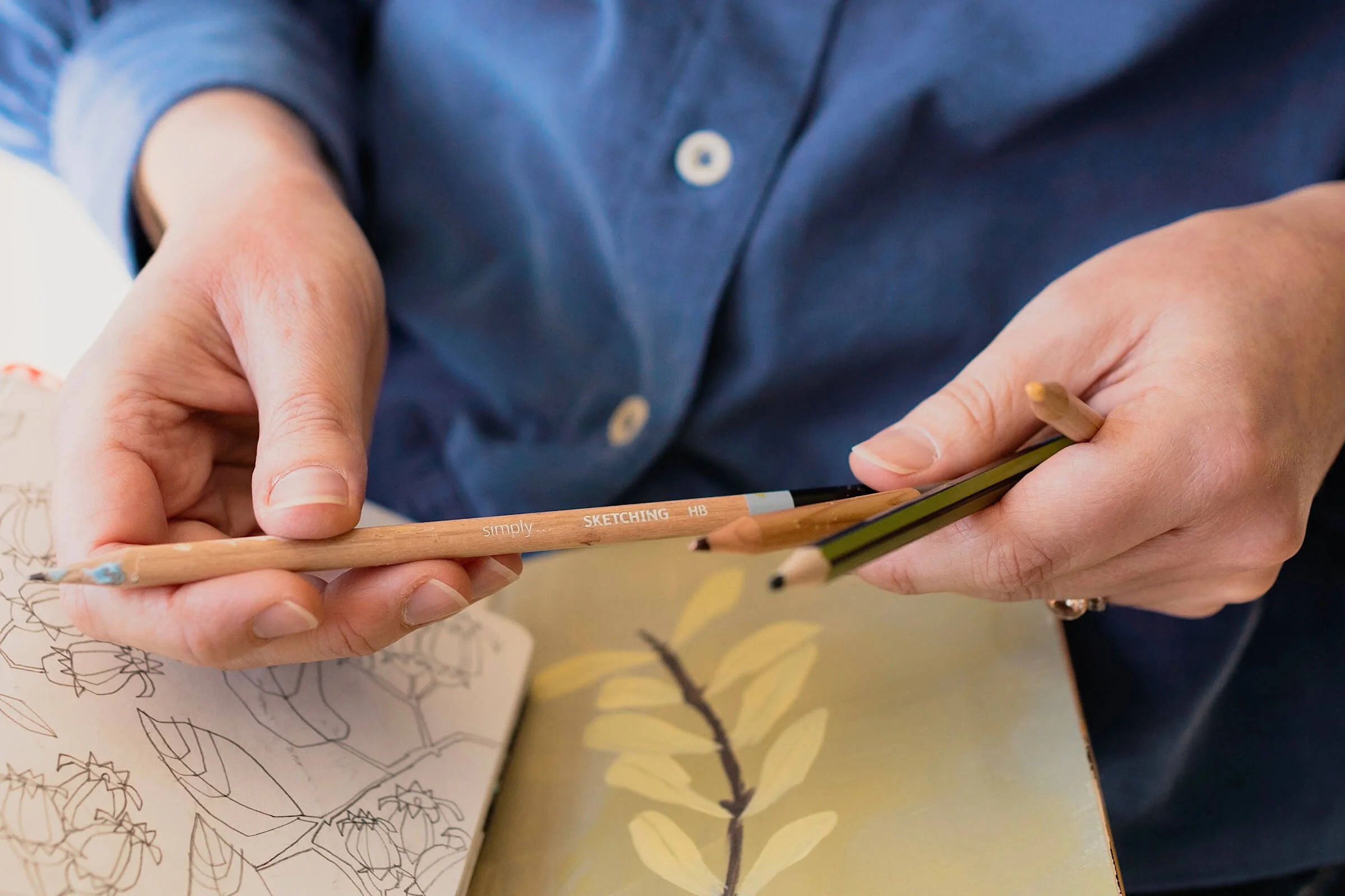 Close-up of artist Ali Mackie's hands holding a selection of graphite sketching and duo-colour pencils over an open sketchbook. The sketchbook reveals a loose botanical drawing, capturing a mindful moment from the contemporary drawing workshop.