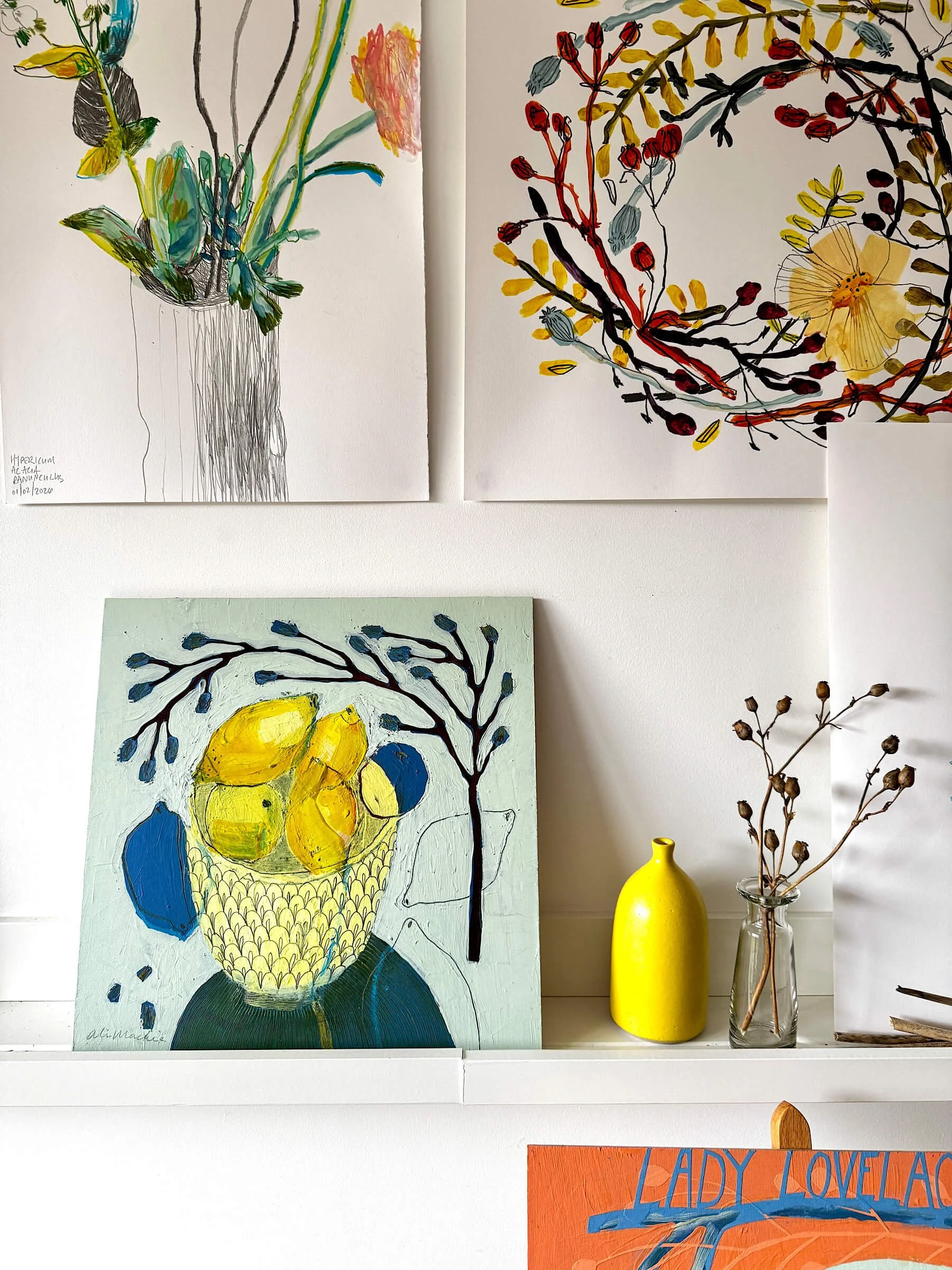 A square contemporary painting of yellow lemons in a patterned bowl on a light blue background is displayed on a white studio shelf next to a bright yellow vase and a glass bottle with dried seed pods.