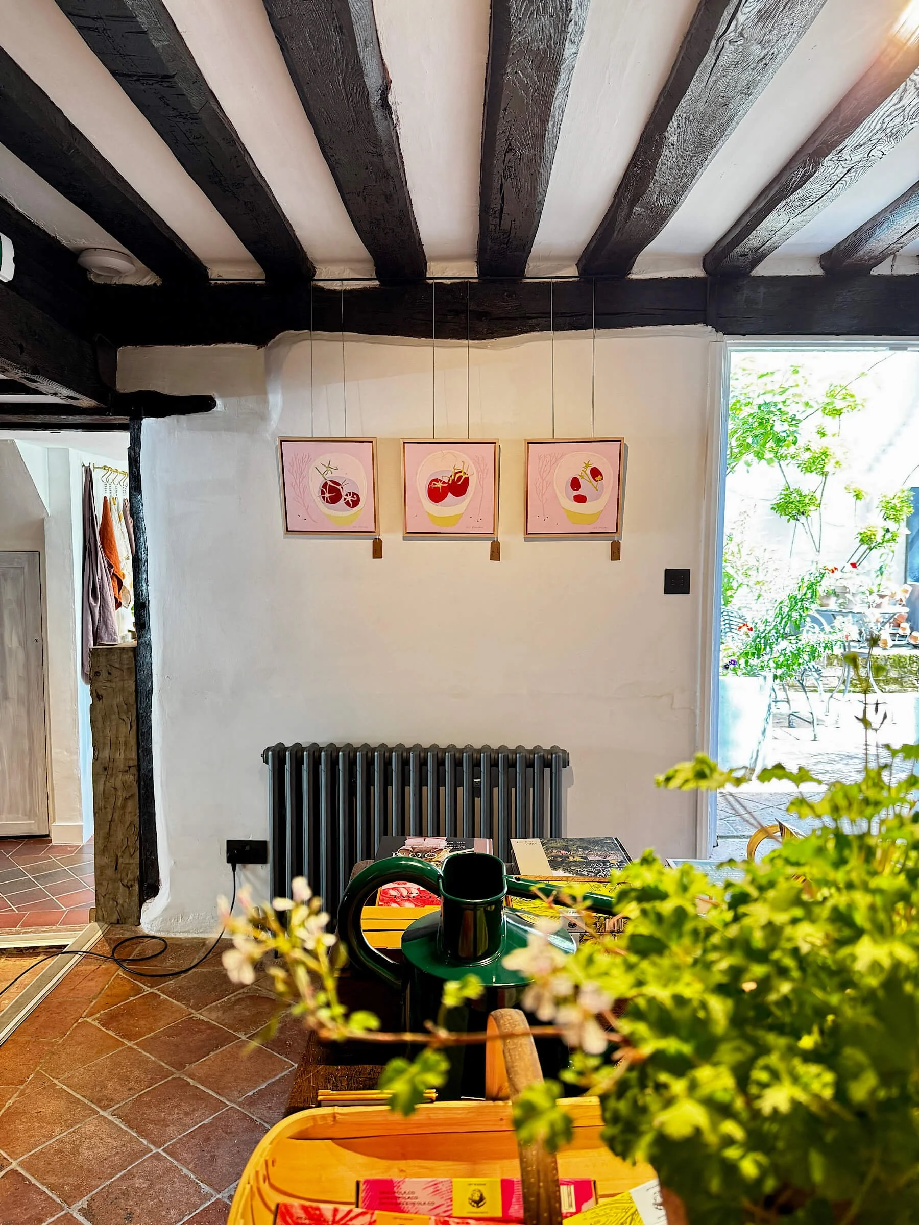 Three framed contemporary still life paintings on board featuring red tomatoes on a pink background, hanging in a row on a white wall beneath historic dark wooden ceiling beams at The Folde gallery.