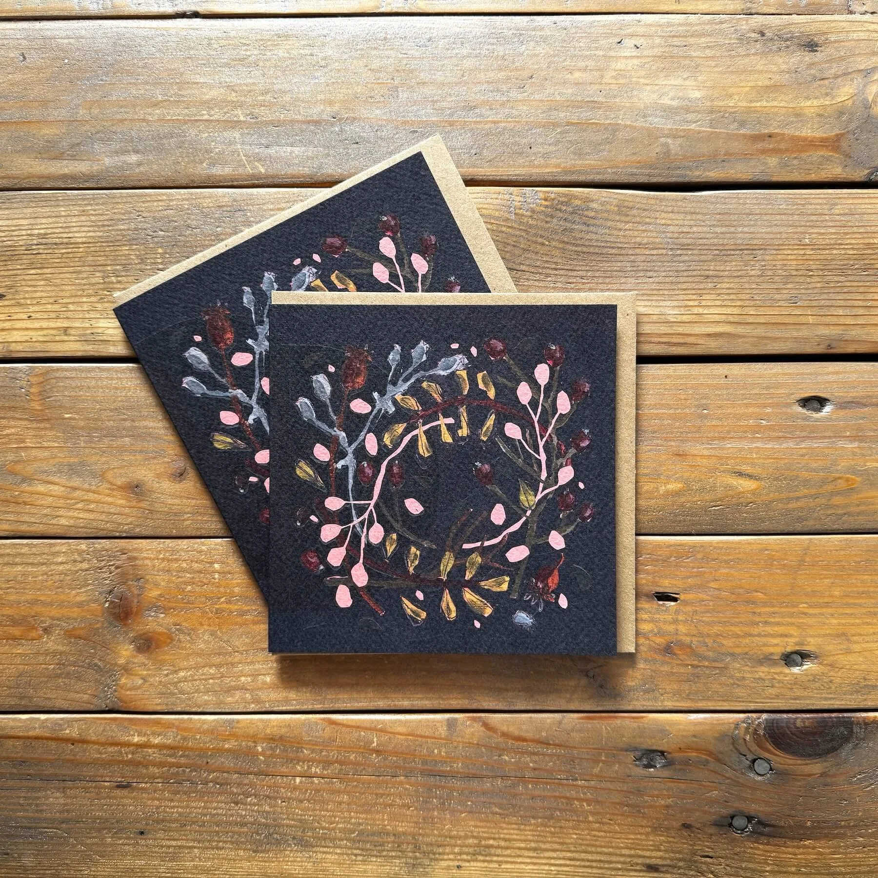 Pair of dark navy abstract botanical wreath Christmas cards with pink buds and gold foliage accents on rustic kraft envelopes.