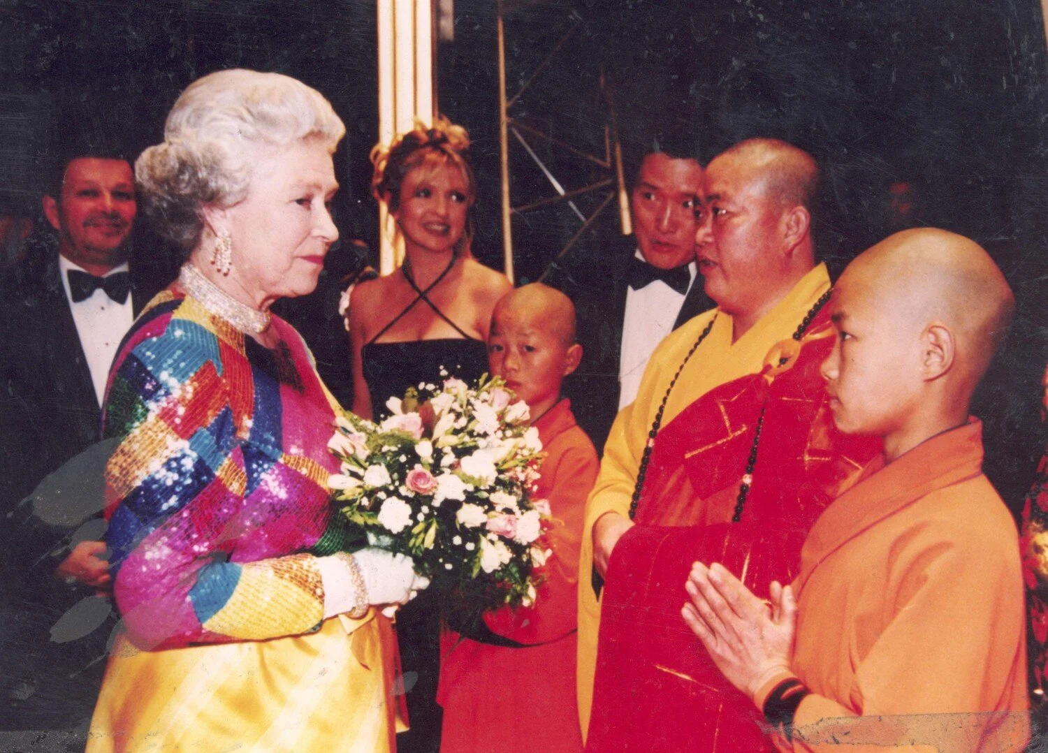 Queen Elizabeth II presenting a bouquet to a Buddhist monk during a formal event, with several other individuals dressed in formal attire and traditional clothing.