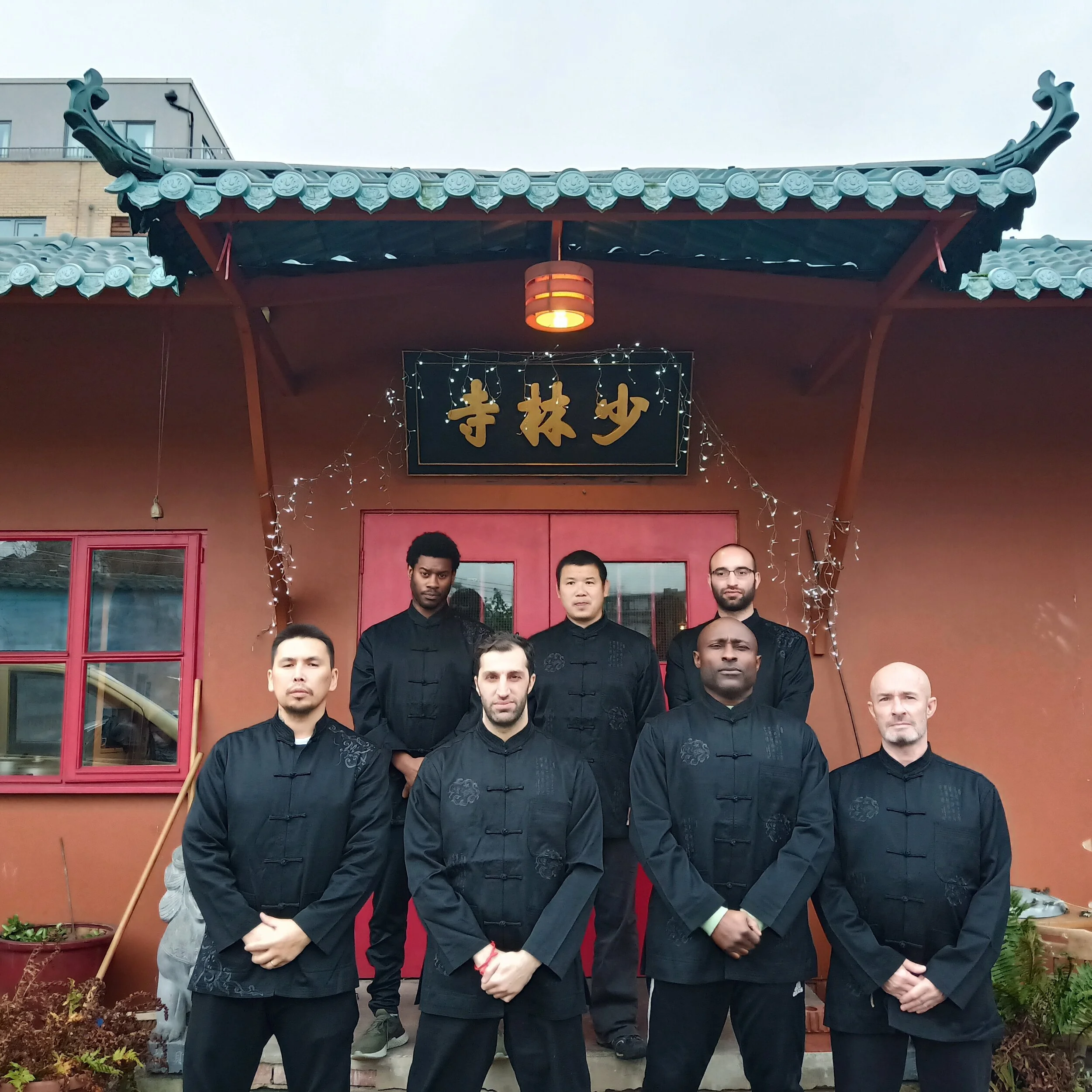Group of six men in traditional black martial arts uniforms standing in front of a building with Asian architectural features and a black sign with gold Chinese characters, next to a window and plants.