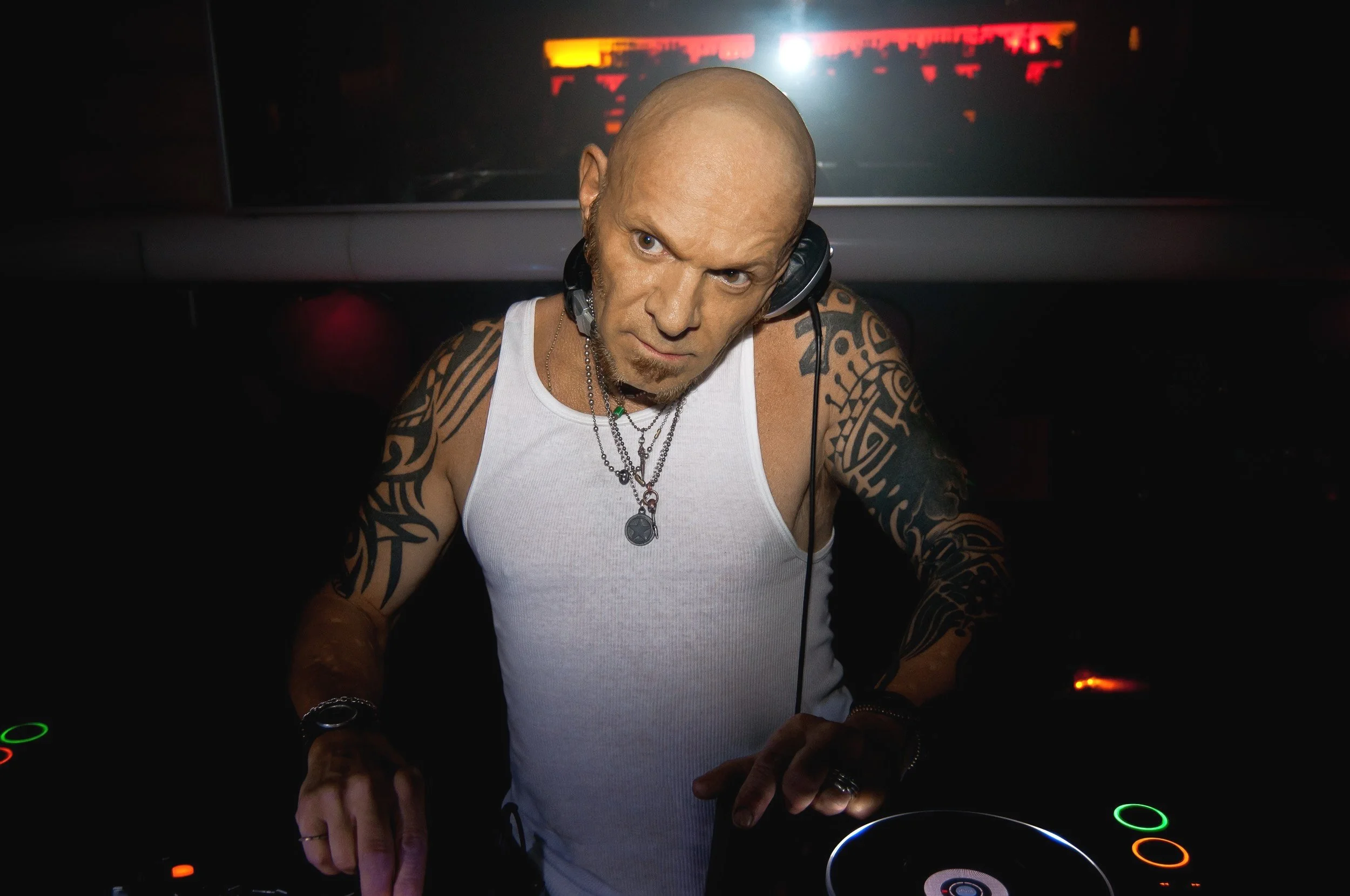 A tattooed male DJ wearing a white tank top and headphones is performing at a dark nightclub, with a blurred DJ equipment in front of him and a faint colorful background projection.