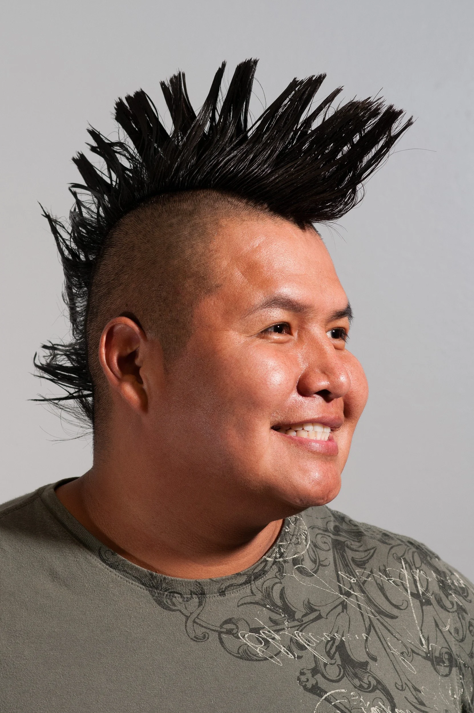Man with a mohawk hairstyle, with the sides of his head shaved close, smiling and wearing a gray T-shirt with black decorative patterns.