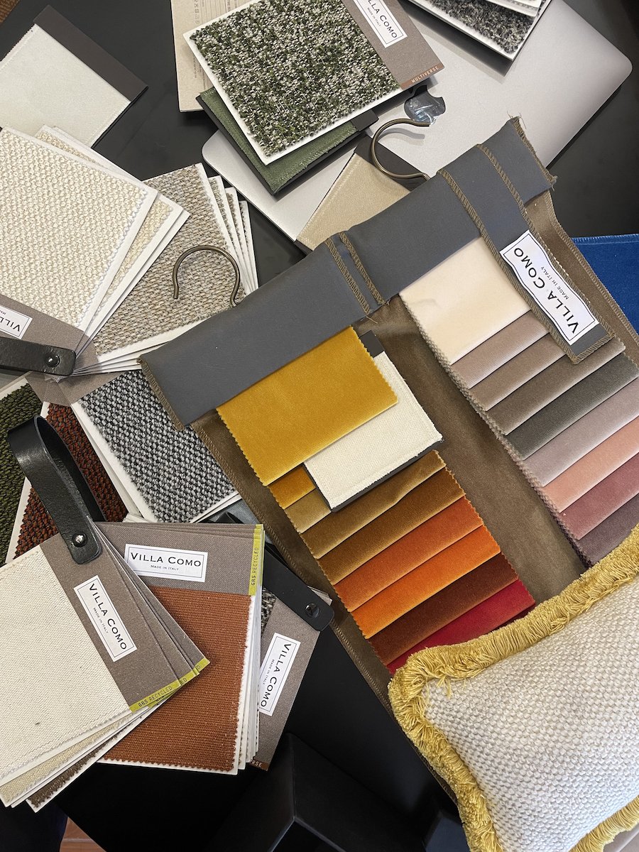 Fabric swatches, color samples, and carpet samples arranged on a table, branded Villa Como, with some samples attached to hooks and others laid flat. Villa Como