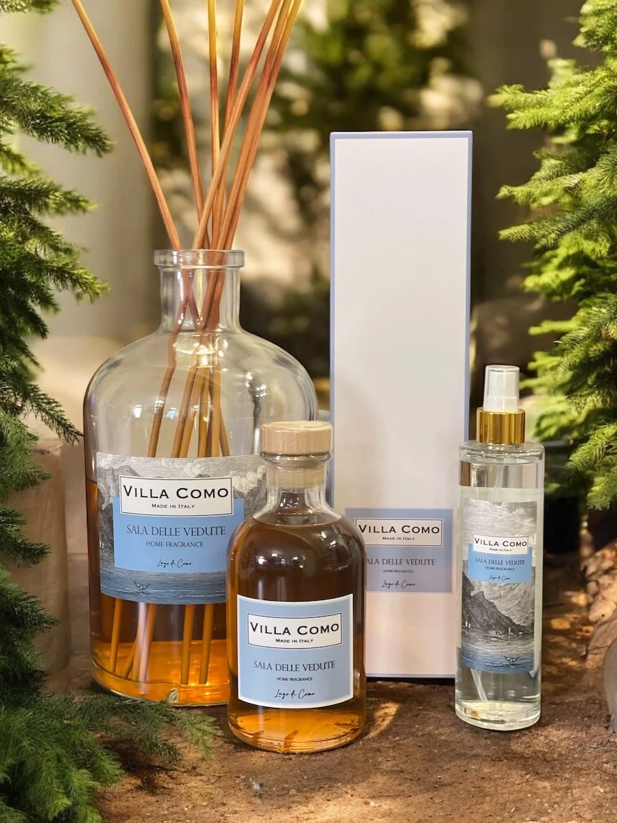 Three Villa Como home fragrance bottles, one large glass jar with reed diffusers, one smaller glass bottle, and one tall spray bottle, placed on a wooden surface with green foliage on sides. Villa Como Home Fragrance