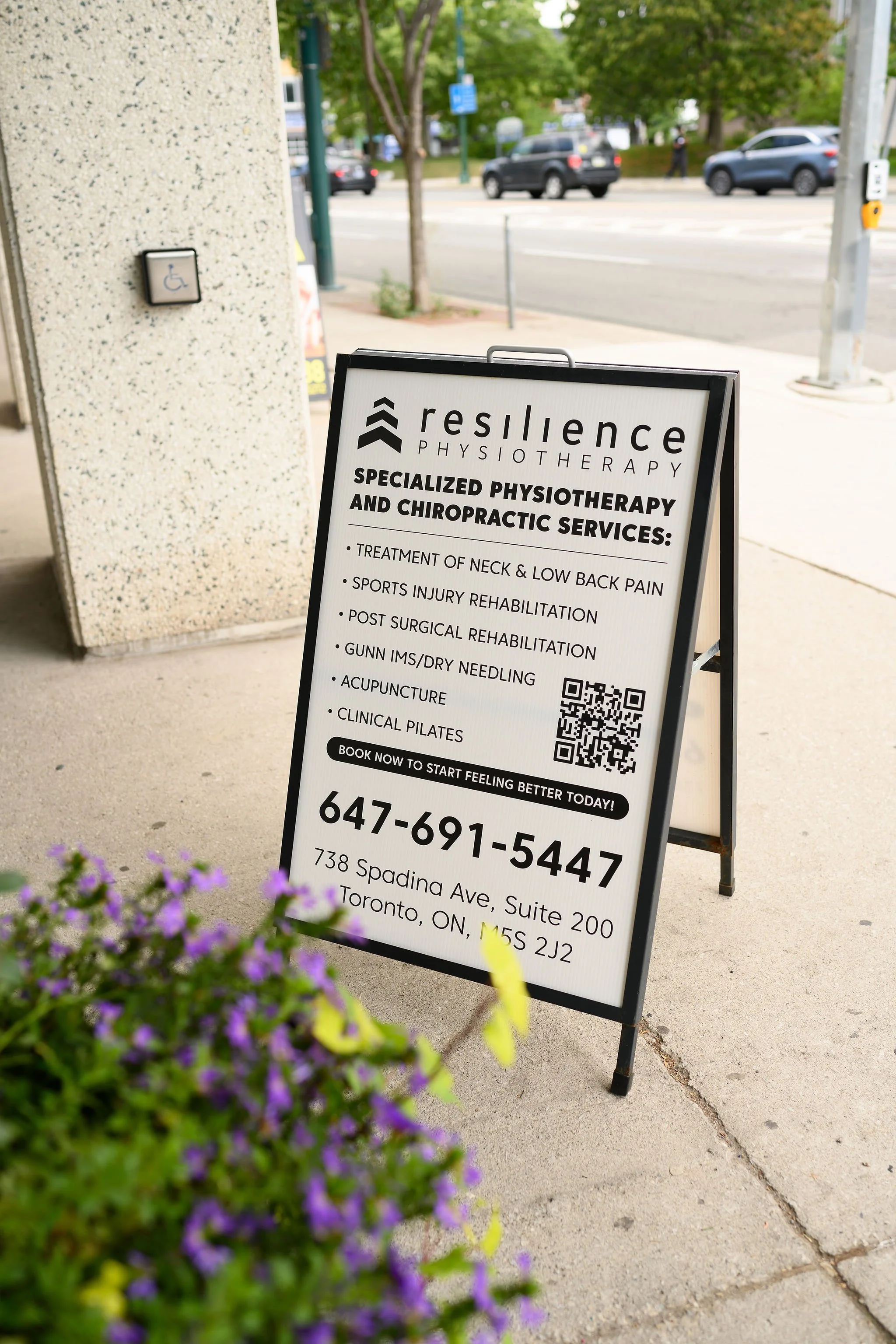 An outdoor sidewalk sign advertising Resilience Physiotherapy with details about services, contact info, and address in Toronto, Canada. There are cars and trees in the background, and some purple flowers in the foreground.