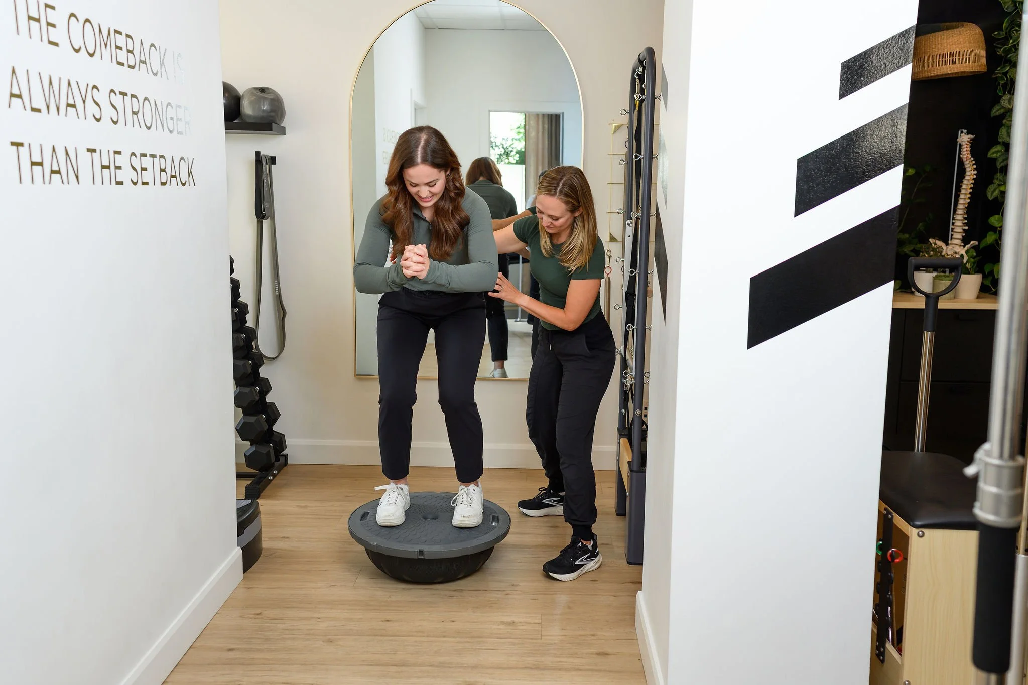 A woman is standing on a balance plate on a stability trainer, smiling and holding her hands together, while a trainer guides her from behind in a gym with equipment and wall decor.