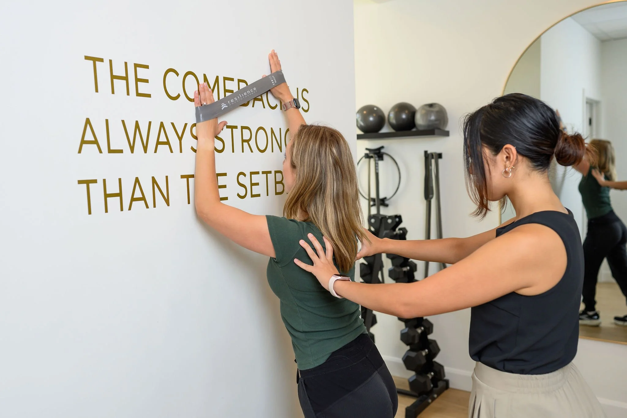 A woman is stretching a band between her hands against a wall. She is being assisted by a female physiotherapist. In the background, there are exercise equipment such as black exercise balls and dumbbells.