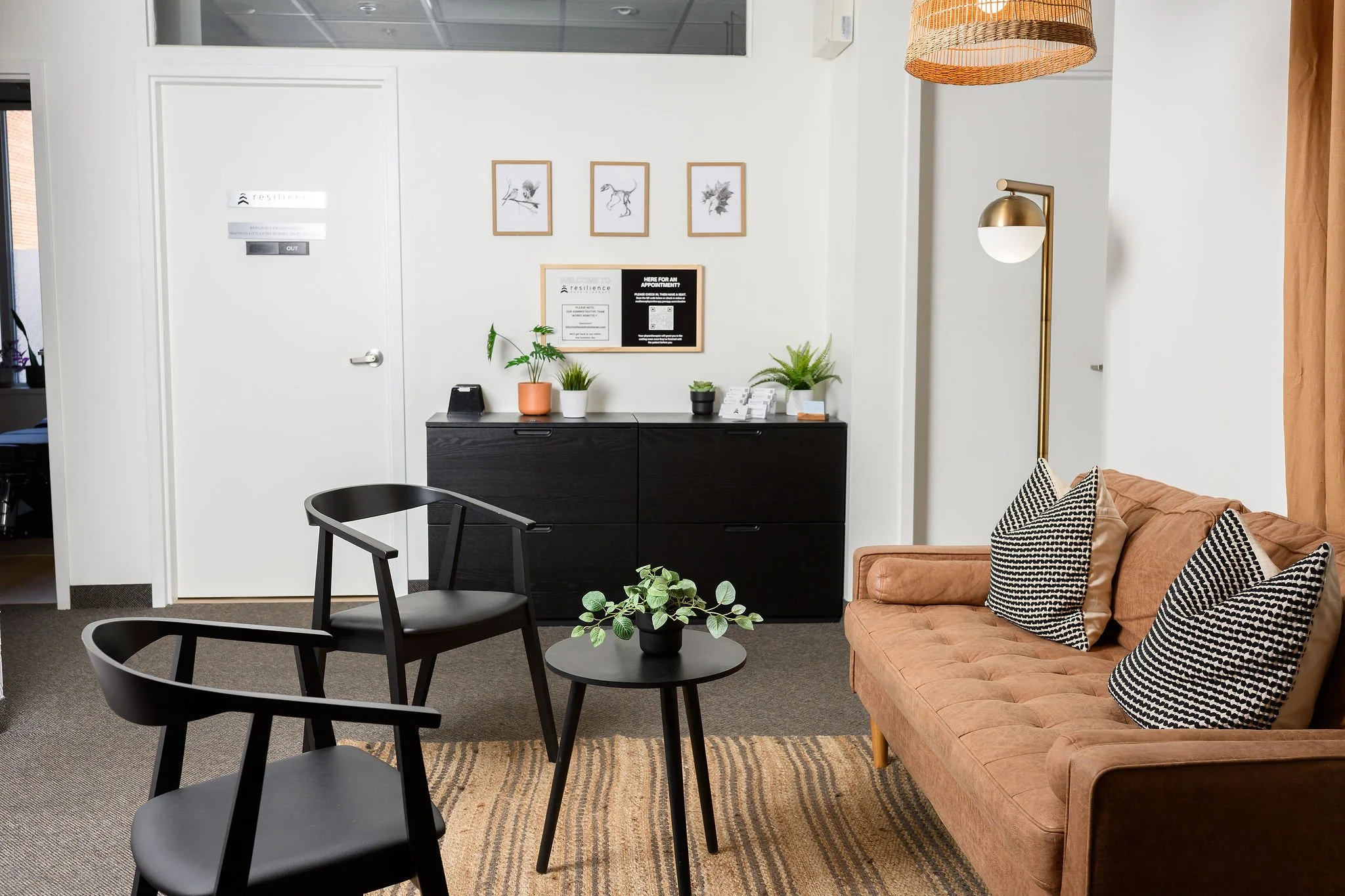 A waiting room features a peach-colored sofa with black and white patterned pillows, a black cabinet topped with potted plants, and framed botanical artwork on a white wall. There are black chairs with circular seats and a small round table with a po