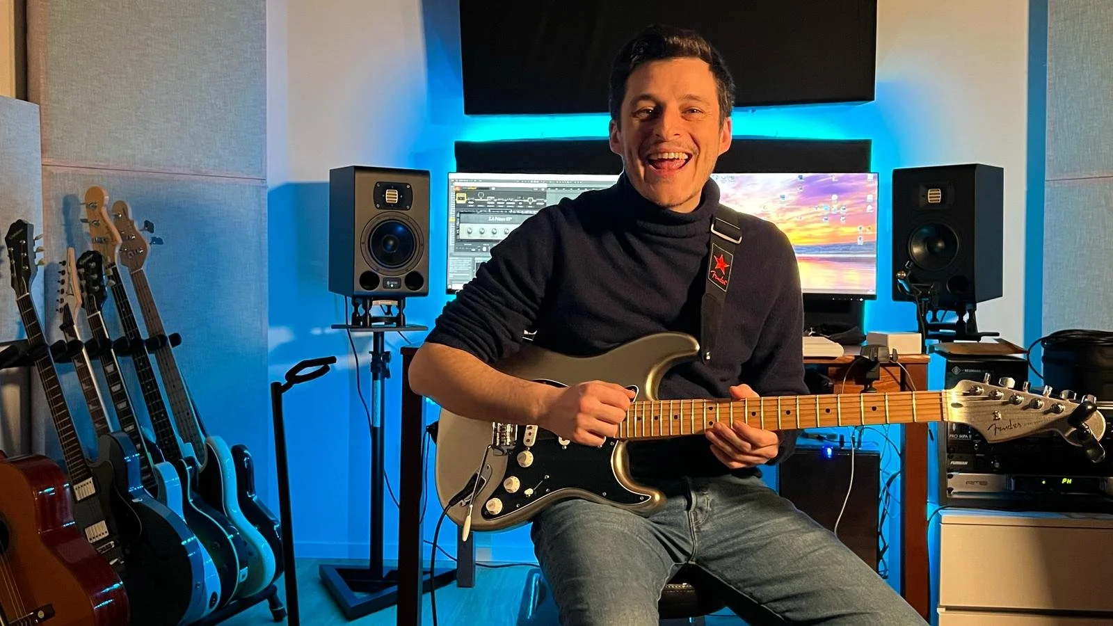 Mike Sorrentino sitting in his music studio, playing an electric guitar, surrounded by multiple guitars, studio monitors, and computer screens with audio editing software, illuminated by blue lighting.