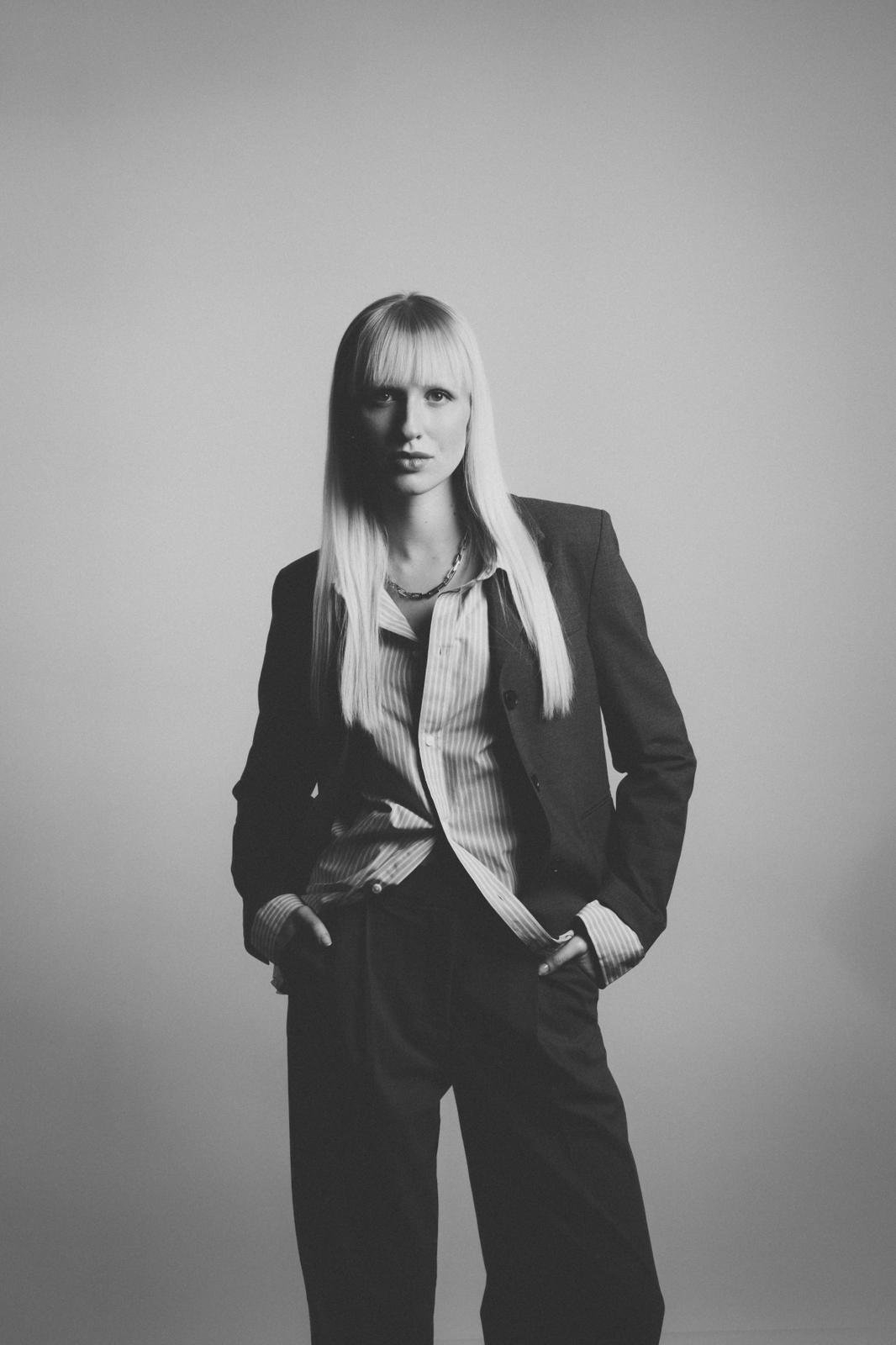 Schwarz-Weiß-Fotografie einer stilvollen Frau mit langen blonden Haaren, in Anzug gekleidet, vor neutralem Hintergrund.