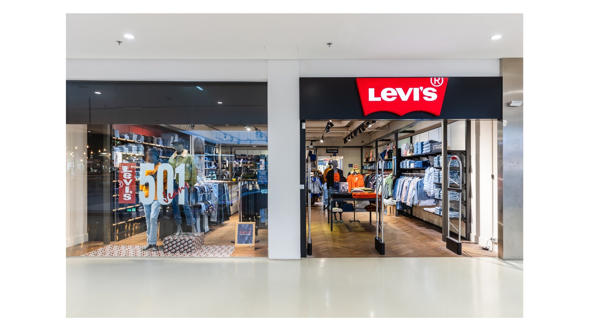 Eingang eines Levi's Bekleidungsgeschäfts mit großen Levis-Logo an der Fassade, im Schaufenster hängen Kleidungsstücke und es gibt Sale-Schilder, die Rabattaktionen ankündigen.