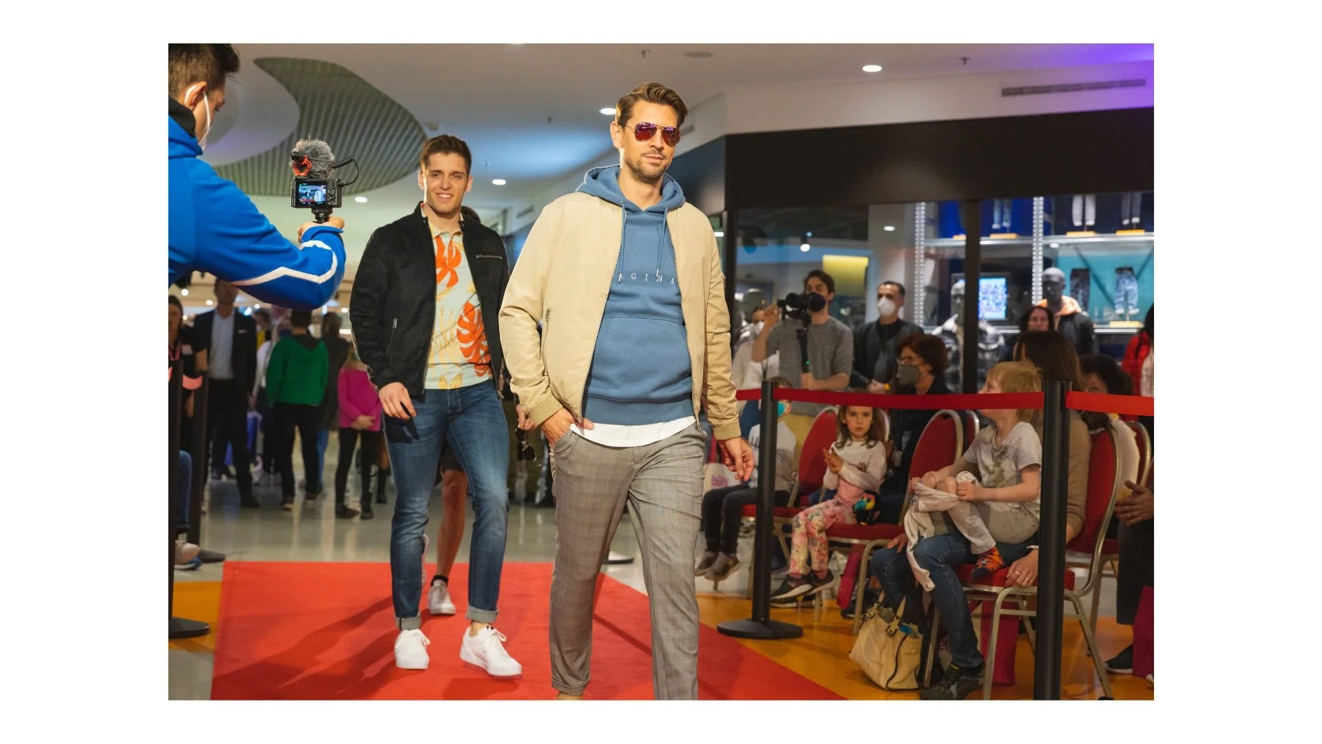 Männer laufen auf einer roten Matte durch eine geschmückte Halle, während eine Person mit Kamera sie filmt. Es sitzen Kinder und Erwachsene im Publikum, manche tragen Masken.