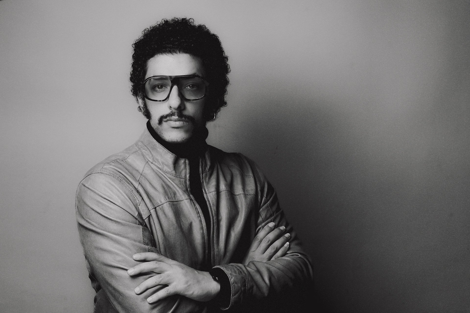 Schwarz-weiß Porträt eines Mannes mit lockigem Haar und Schnurrbart, trägt große Brille und Lederjacke, steht mit verschränkten Armen vor einer neutralen Wand.