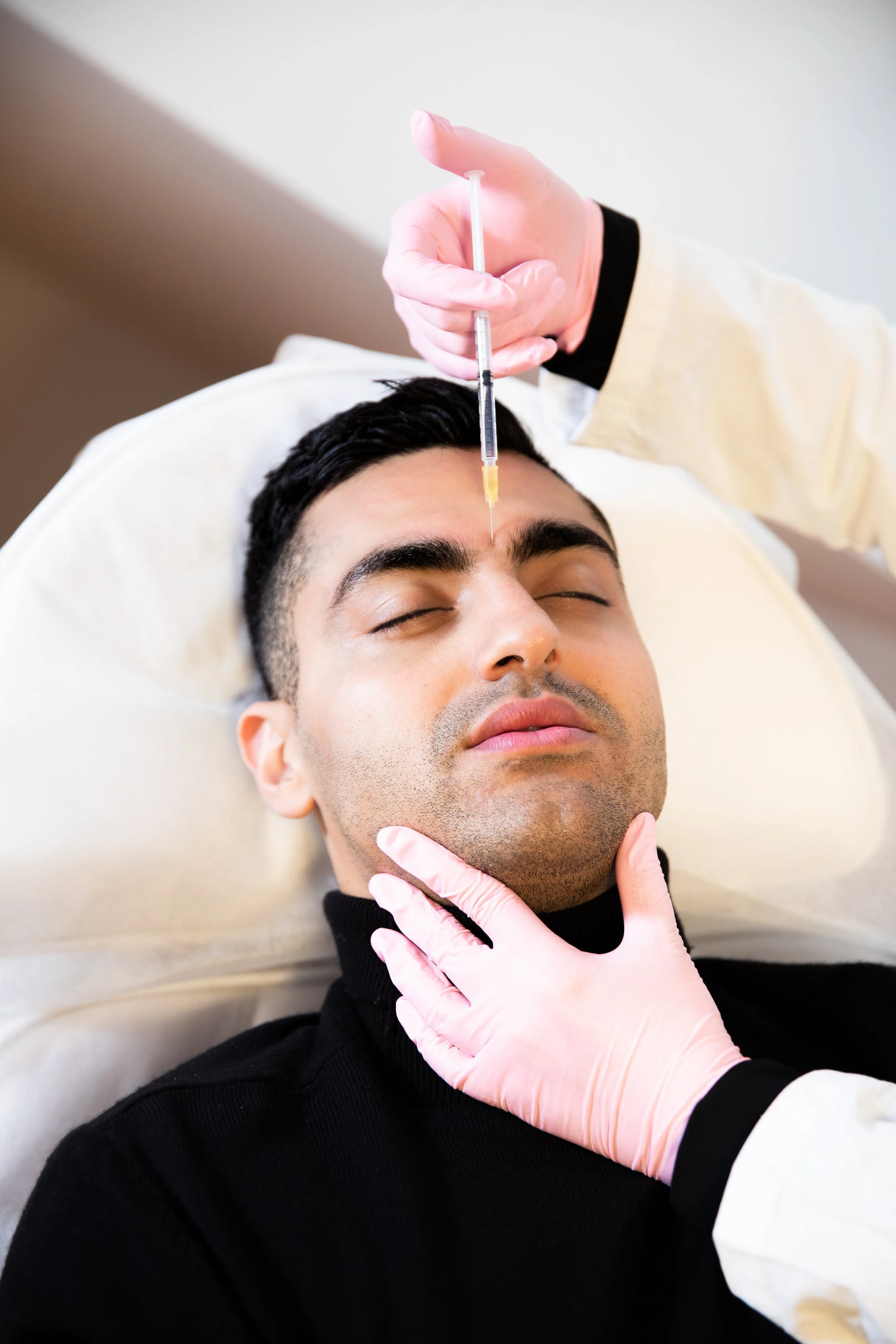 Ein Mann liegt entspannt mit geschlossenen Augen auf einer Behandlungsliege, während eine medizinische Fachkraft ihm eine Injektion in die Stirn verabreicht.