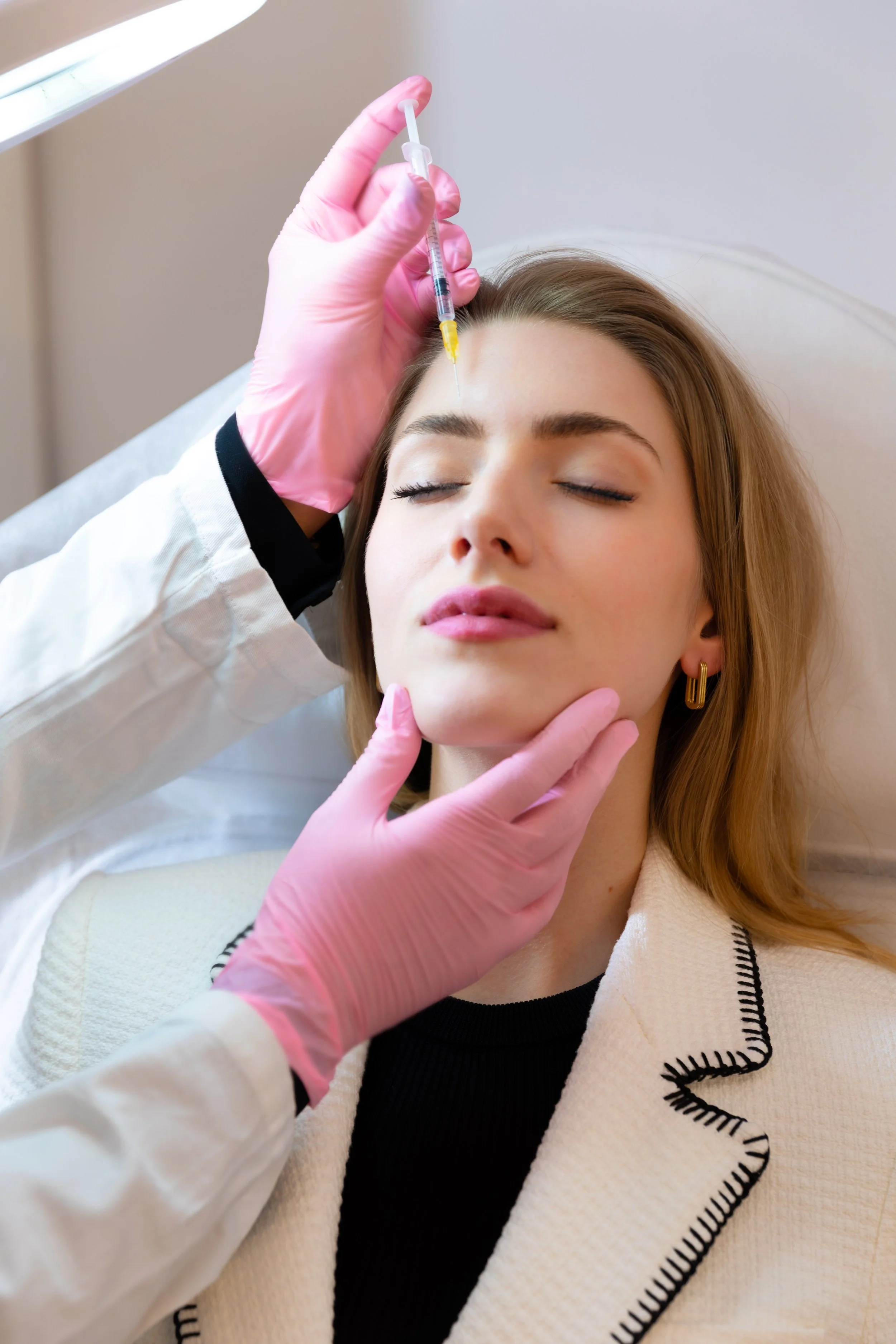 Eine junge Frau liegt entspannt auf einer Liege, während eine medizinische Fachkraft ihr eine Injektion in die Stirn gibt, vermutlich Botox oder ähnliche Behandlung.