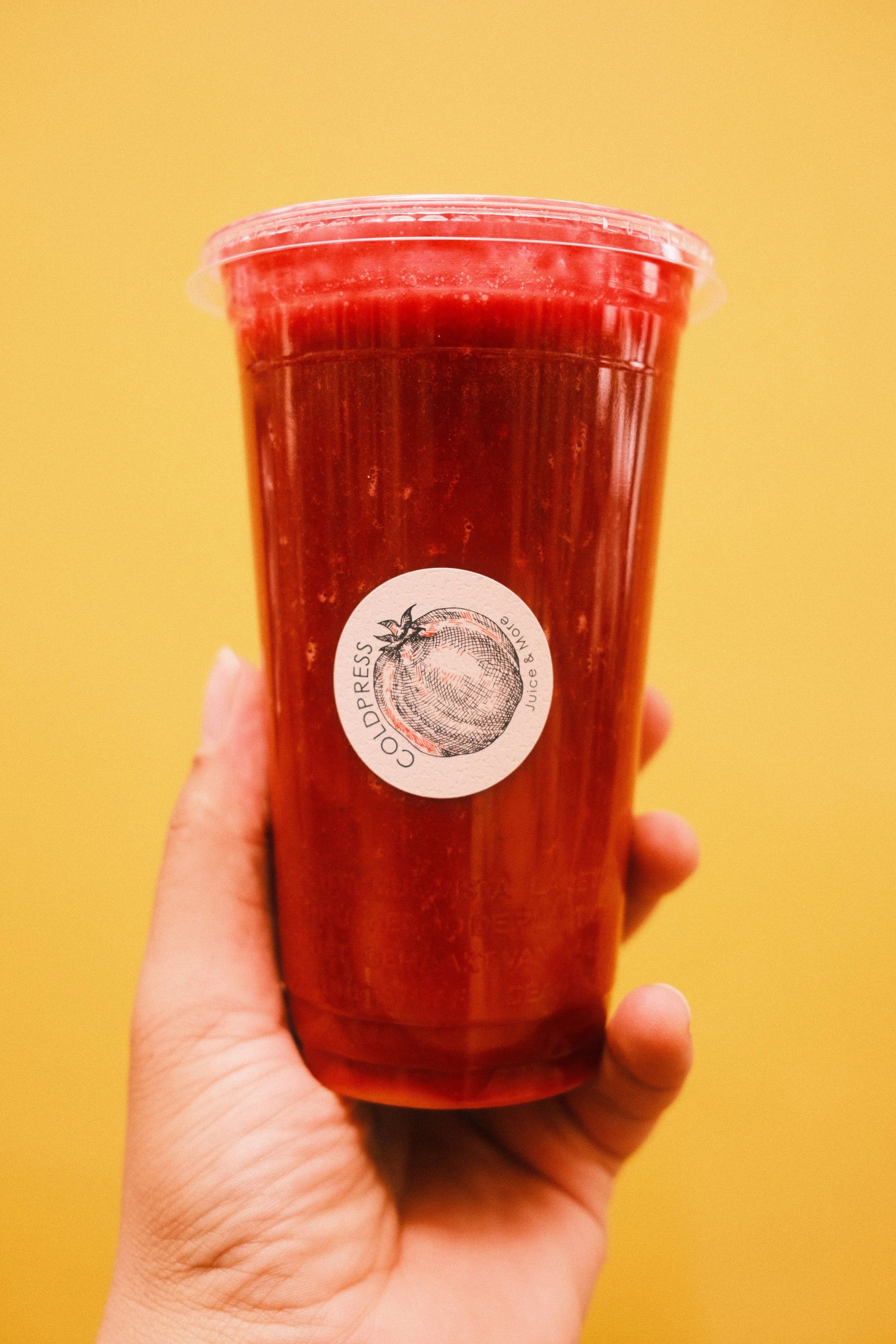 Hand hält einen großen roten Smoothie in einem transparenten Becher mit Deckel, auf dem ein rundes Logo mit einem Gemüsetomaten und der Aufschrift 'CUPRESS Juice & More' ist, vor einem gelben Hintergrund.