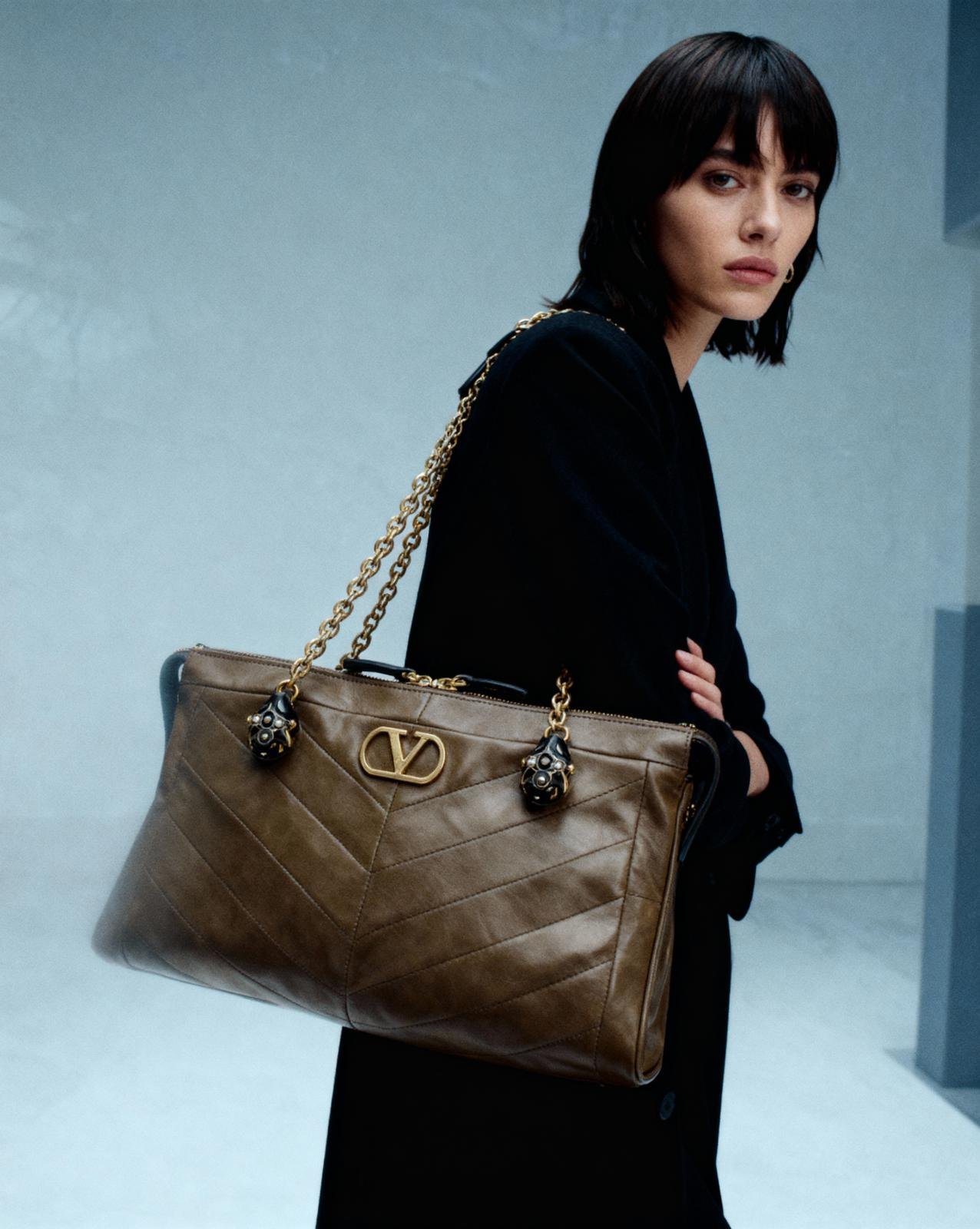 Frau mit schwarzen Haaren trägt eine große braune Designer-Handtasche mit goldener Kette und Logo.