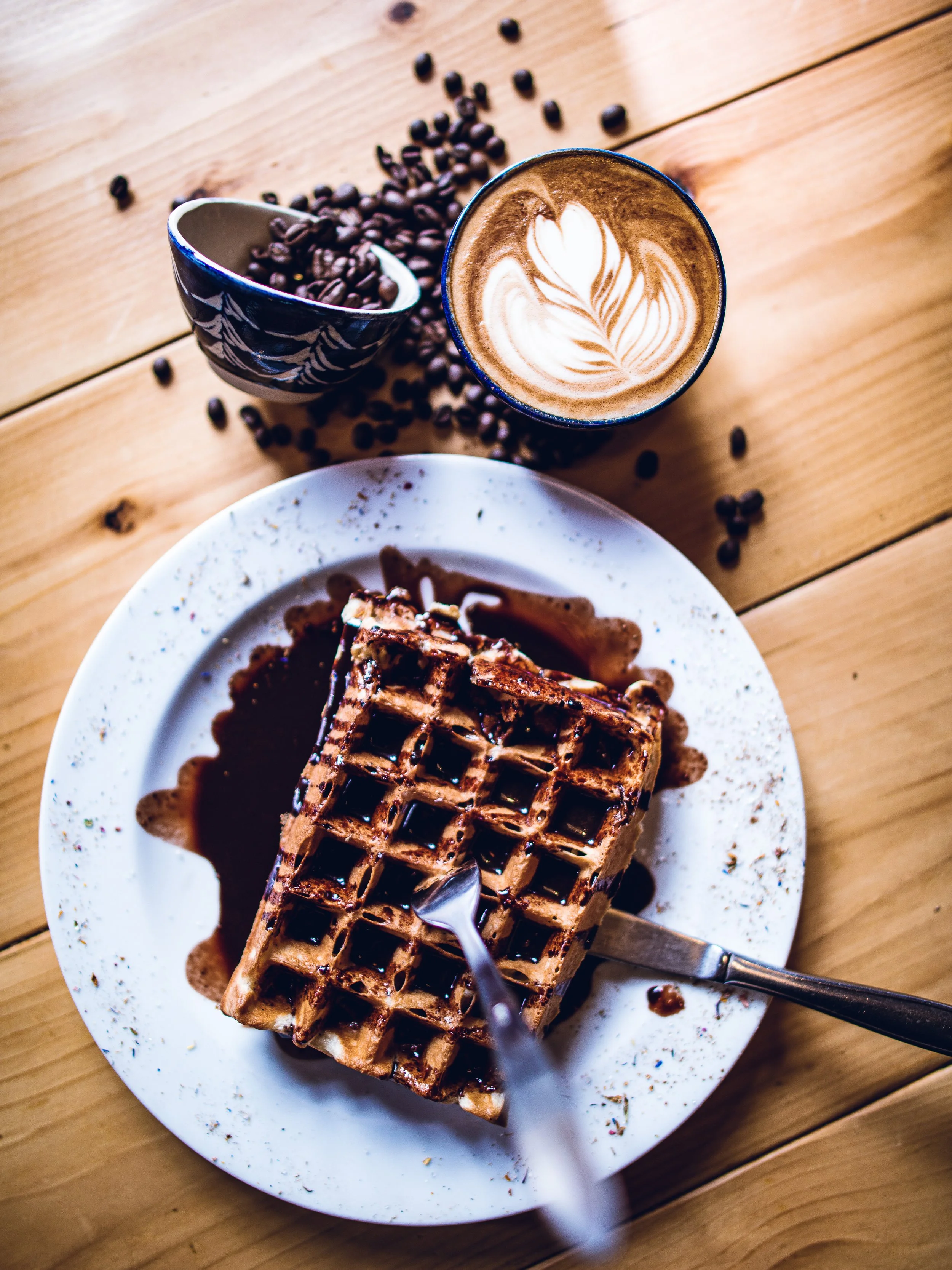 Waffel mit Schokoladensoße, Löffel, Kaffee mit Latte Art, Kaffeebohnen und Kaffeelöffel auf einem Holztisch.