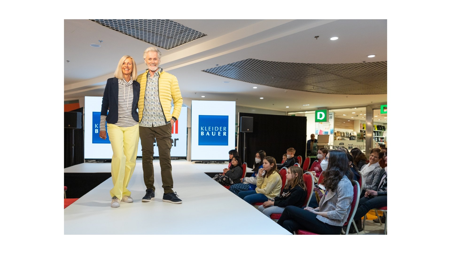 Zwei Menschen stehen auf einer Bühne vor einem Publikum in einem Einkaufszentrum. Es sind verschiedene Werbetafeln mit der Aufschrift 'Kleider Bauer' im Hintergrund sichtbar.