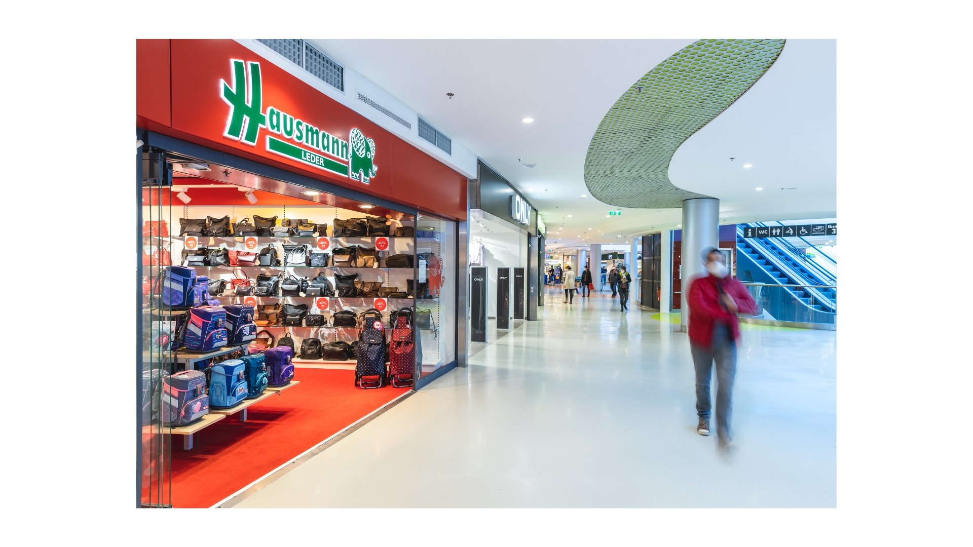 Ein Flughafen-Einkaufsbereich mit einem Geschäft für Taschen und Rucksäcke namens 'Hausmann Leder'. Es gibt weitere Geschäfte und Passanten im Hintergrund, einige tragen Masken.