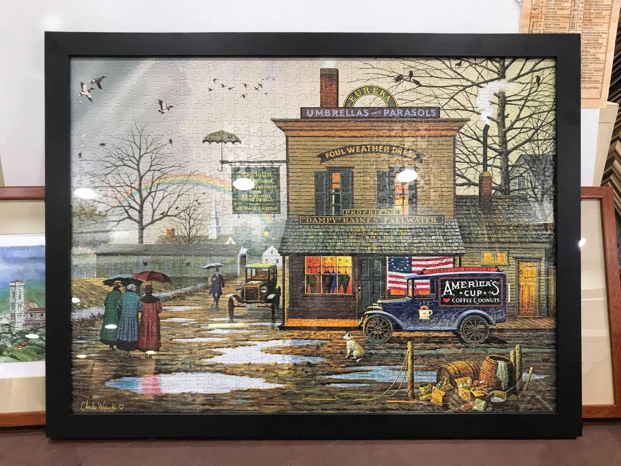 A completed puzzle depicting a vintage scene of a street corner with a brick building labeled "Dampy" Raines, featuring signs for umbrellas and parasols, a fairground-style truck with a sign "America's Cup Coffee & Donuts", and people walking with um