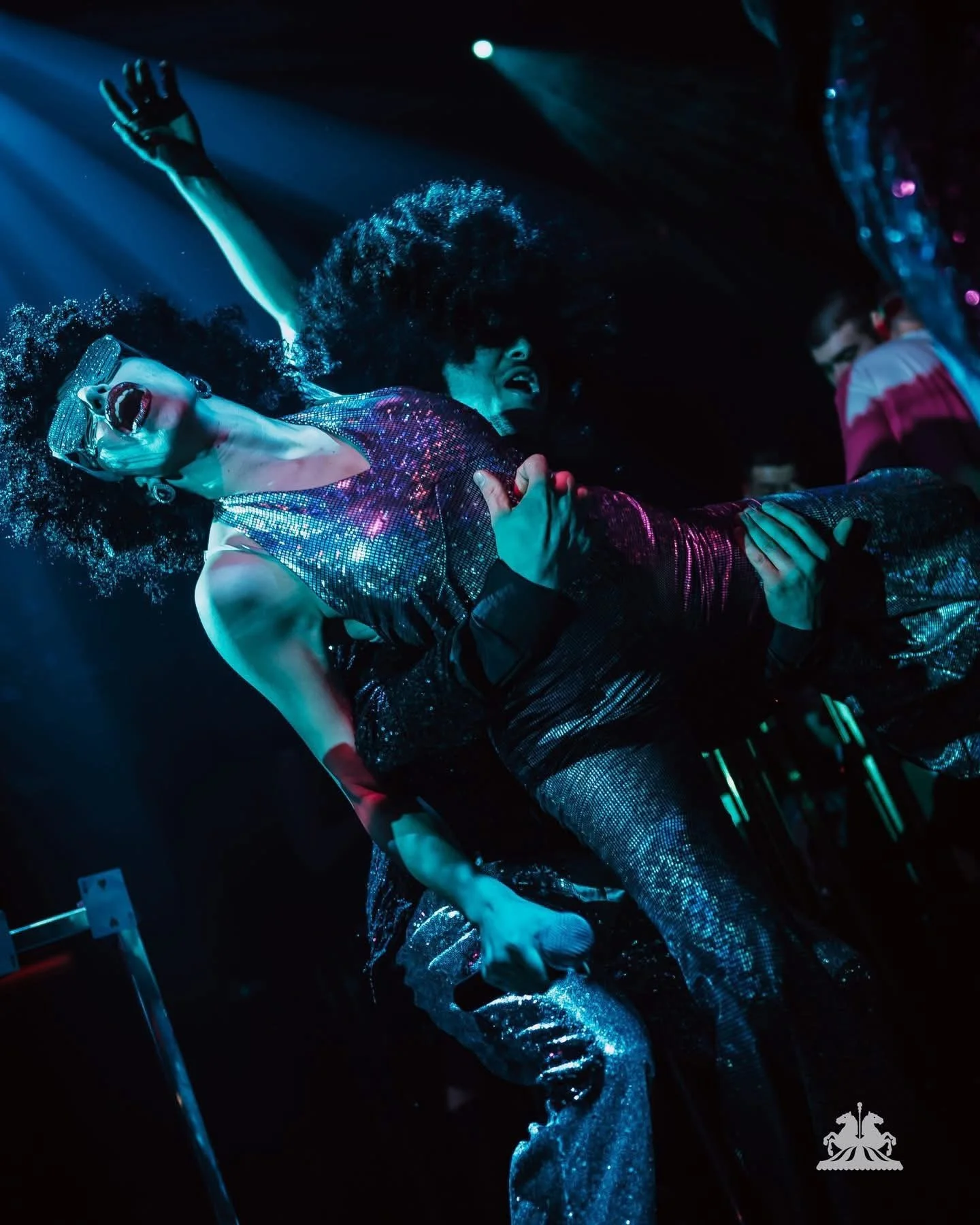 A performer in a shiny, colorful outfit with sunglasses and curly hair, singing passionately on stage with one arm raised, illuminated by blue and purple lights.