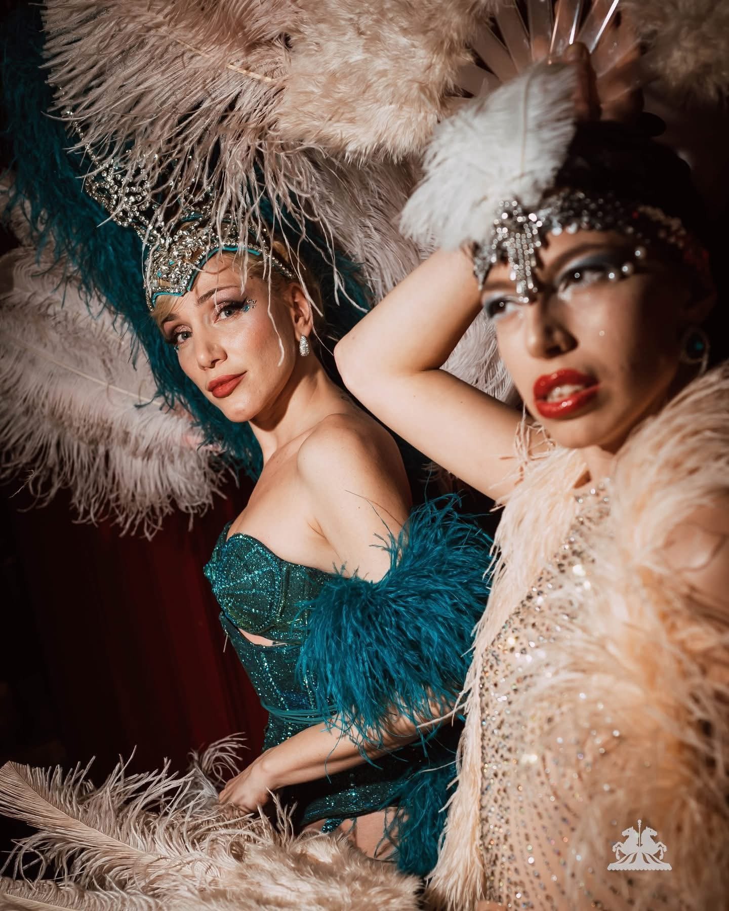 Two women dressed in glamorous costumes with feathered headdresses and sequined dresses, posing together with one smiling and the other looking contemplative, surrounded by feathers.