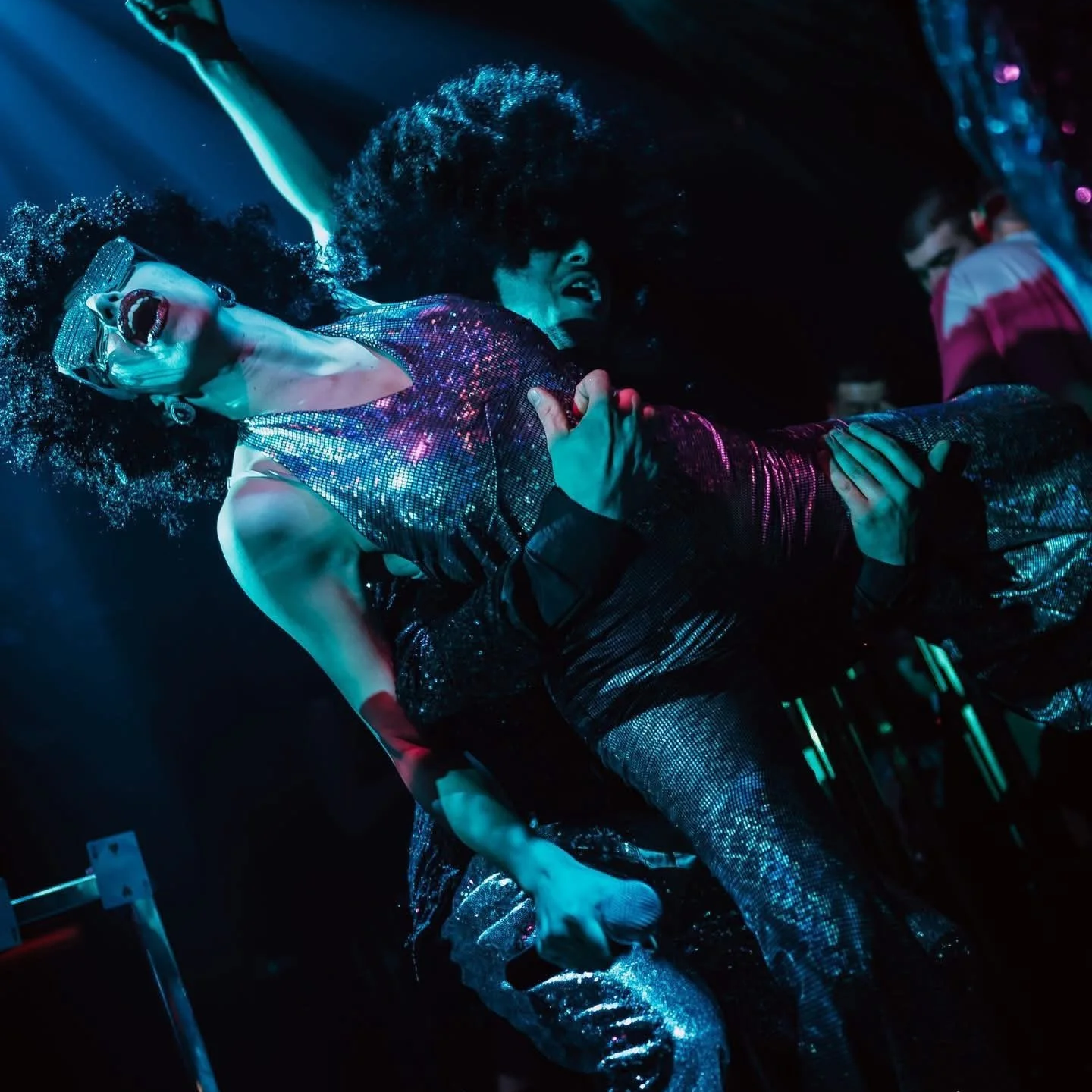Two drag performers on stage with glittering outfits, one showcasing long lipstick and sunglasses, and the other passionately singing or speaking into a microphone, amidst vibrant lighting and energetic atmosphere.