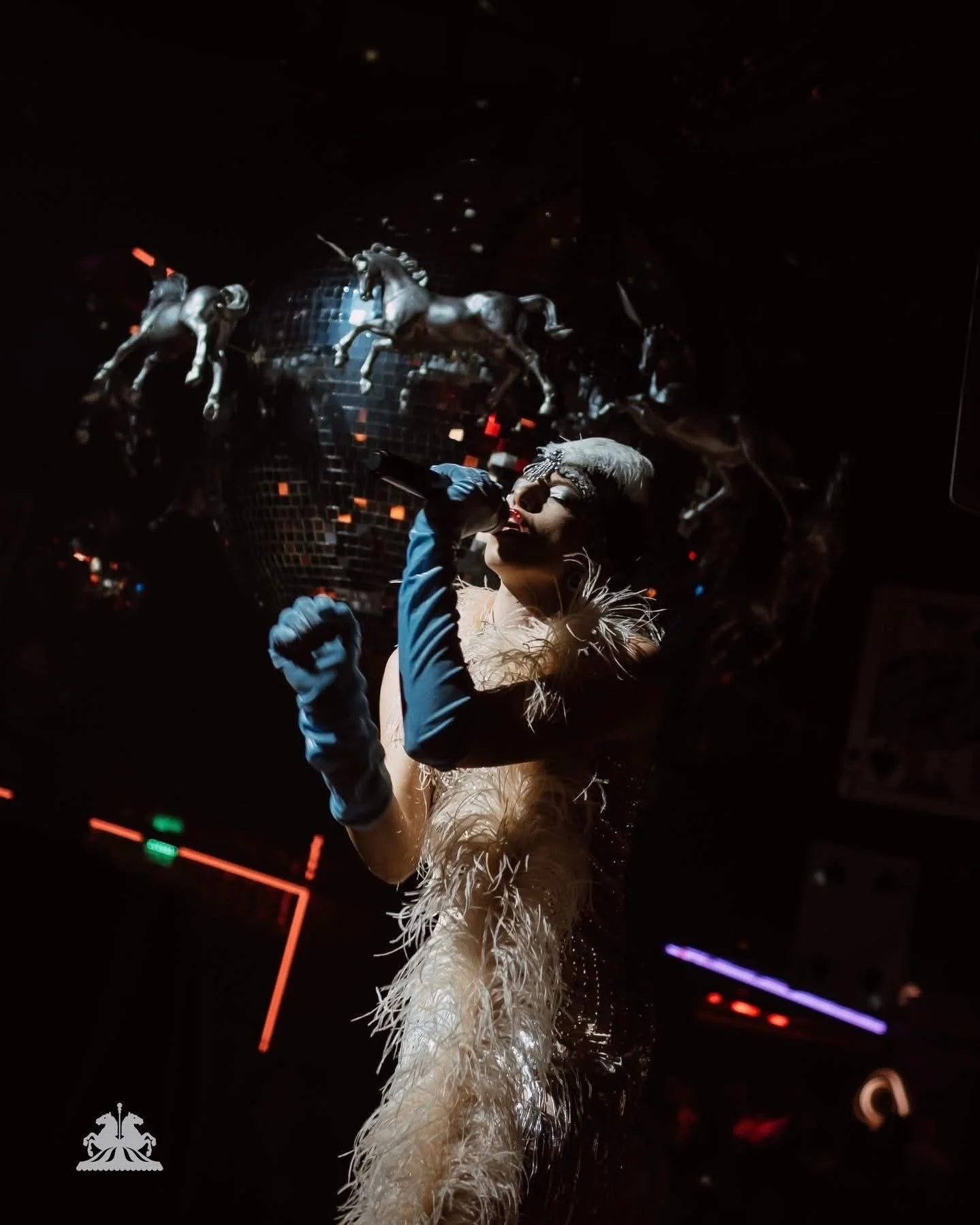 A woman with blonde hair wearing gloves and a feathered costume appears to be singing into a microphone on a dark stage with colorful lights and a disco ball.