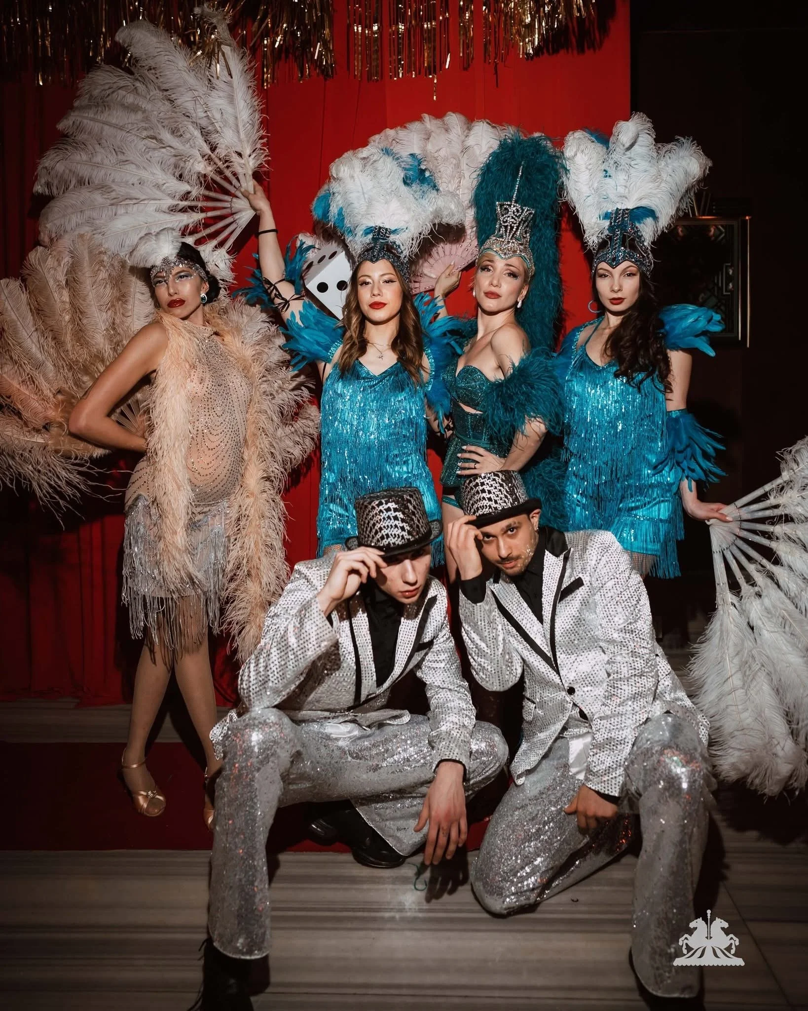 Group of four women dressed in glamorous, sparkling blue outfits with large feathered headpieces, posing in front of a red curtain backdrop at a costume or performance event. Two men in matching black and white costumes with top hats are in the foreg