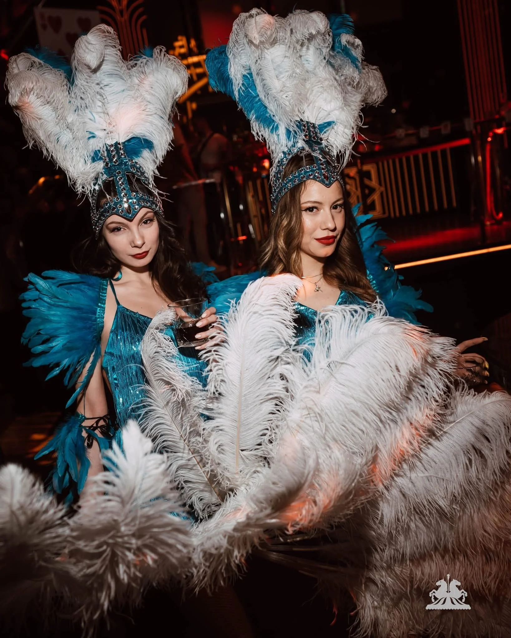 Two women dressed in elaborate show costumes with large feathered headdresses, blue and white feathers, and sparkling embellishments, at a nightclub or stage setting.