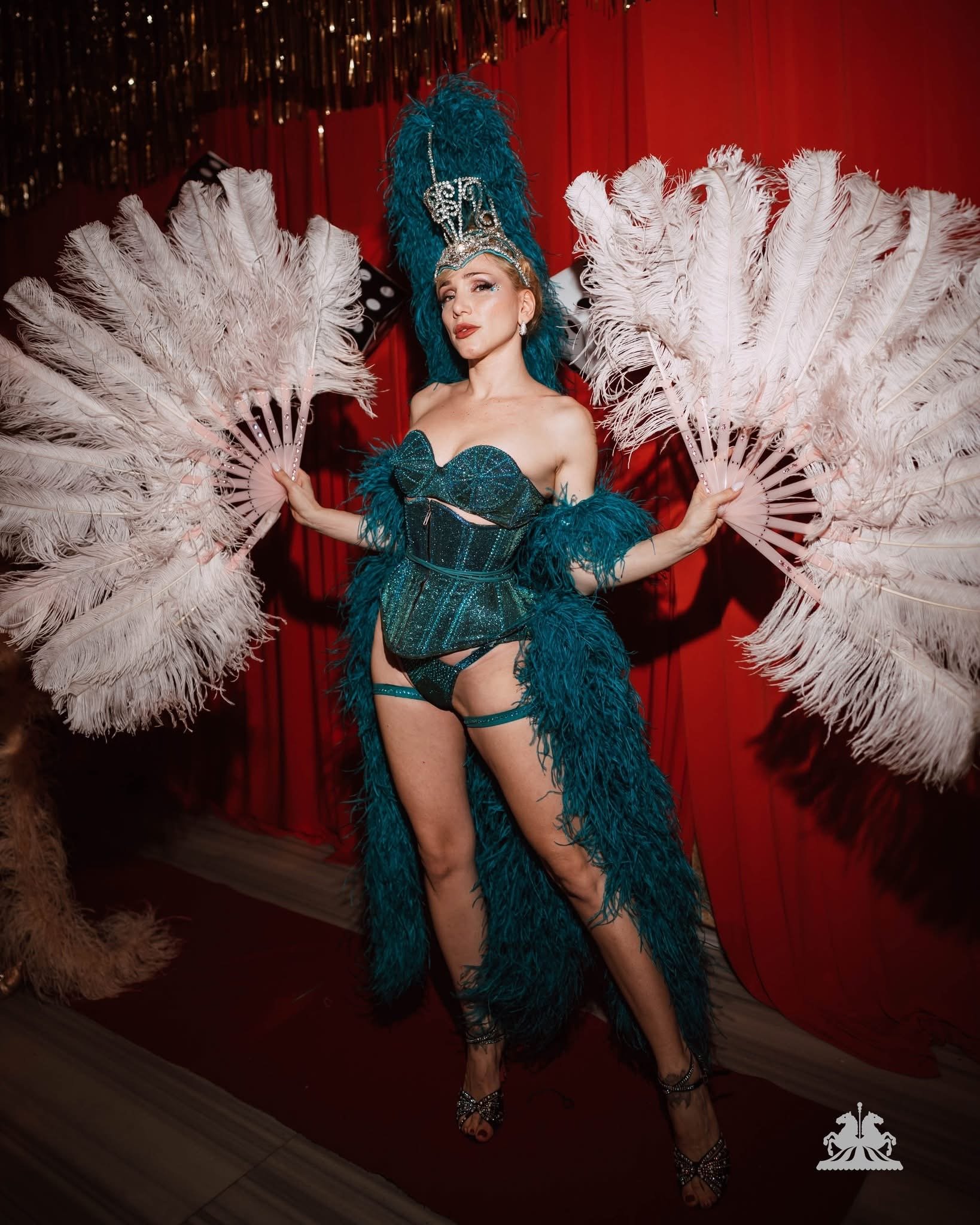 A woman dressed in a sparkling turquoise and blue burlesque costume with a large feathered headdress, holding two large white feather fans, standing in front of a red curtain with gold accents on the ceiling.