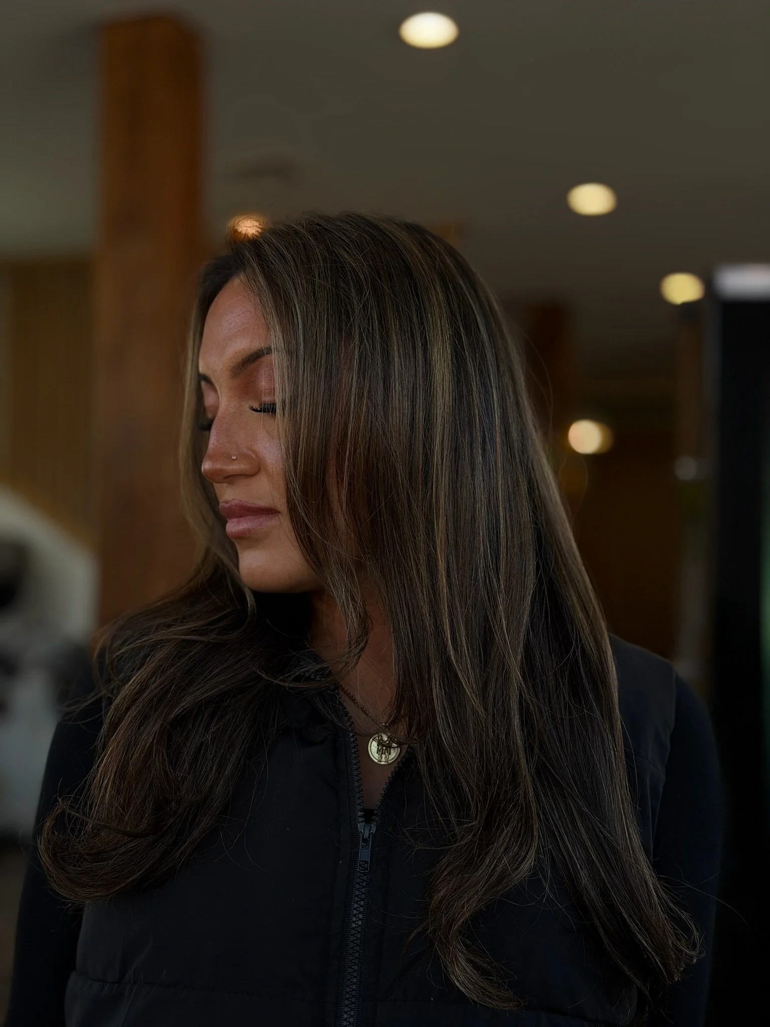 Long dark brown hair with lighter face-framing strands and curved layers, worn over a black zip-front top.