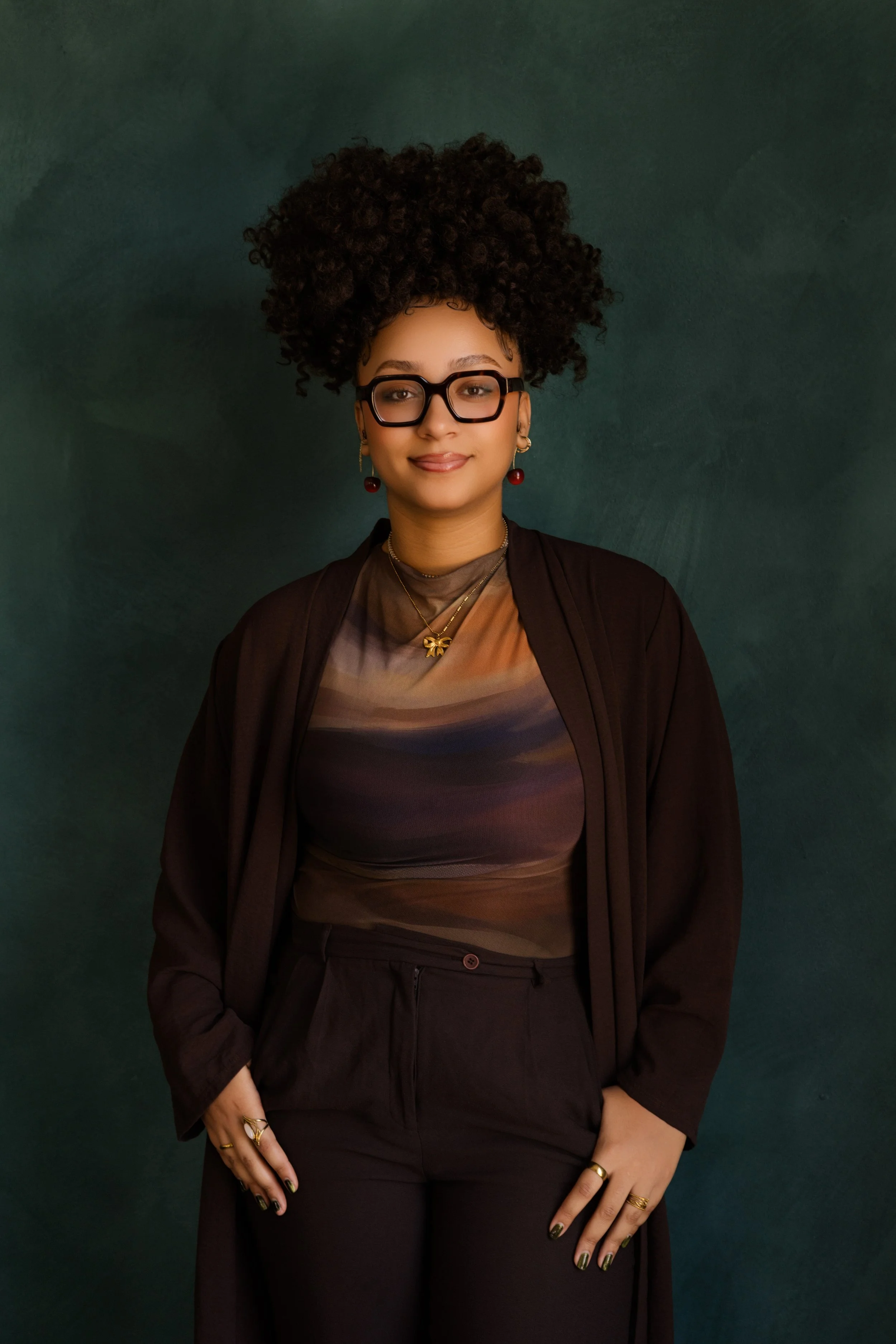 A woman with dark, curly hair styled in a large updo, wearing black-rimmed glasses, earrings, and layered necklaces. She is dressed in a shiny, patterned top and dark pants, standing against a dark green textured background.