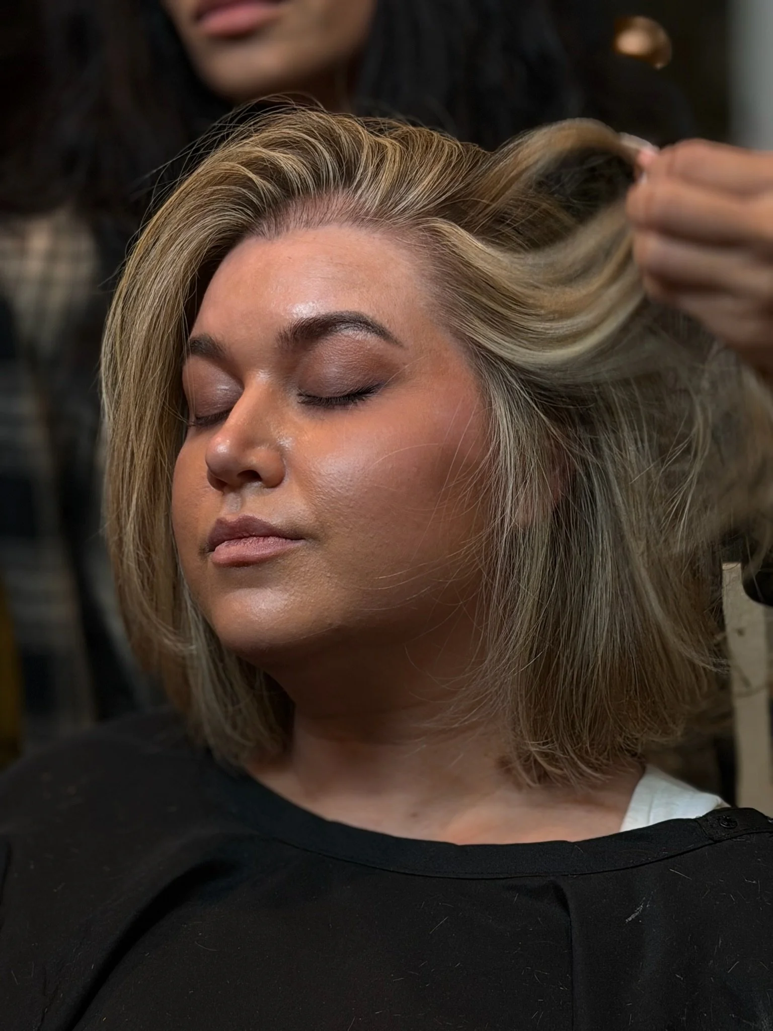 A stylist arranges a client’s short blonde hair; the client wears softly defined eye makeup.