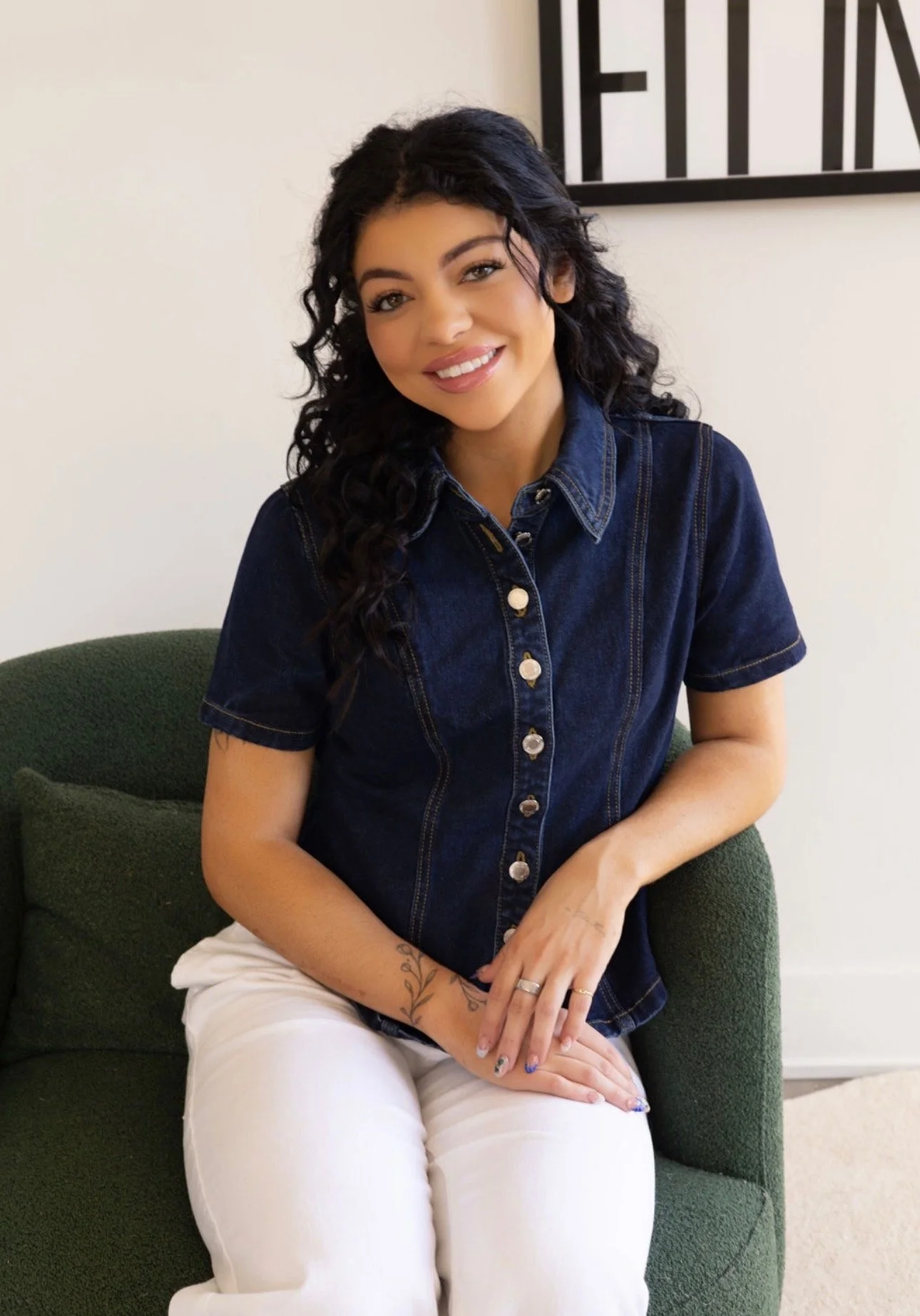 A woman with curly dark hair, smiling, sitting on a green sofa. She is wearing a dark denim shirt and white pants, with tattoos on her left forearm and fingers. There is a framed artwork partially visible on the wall behind her.