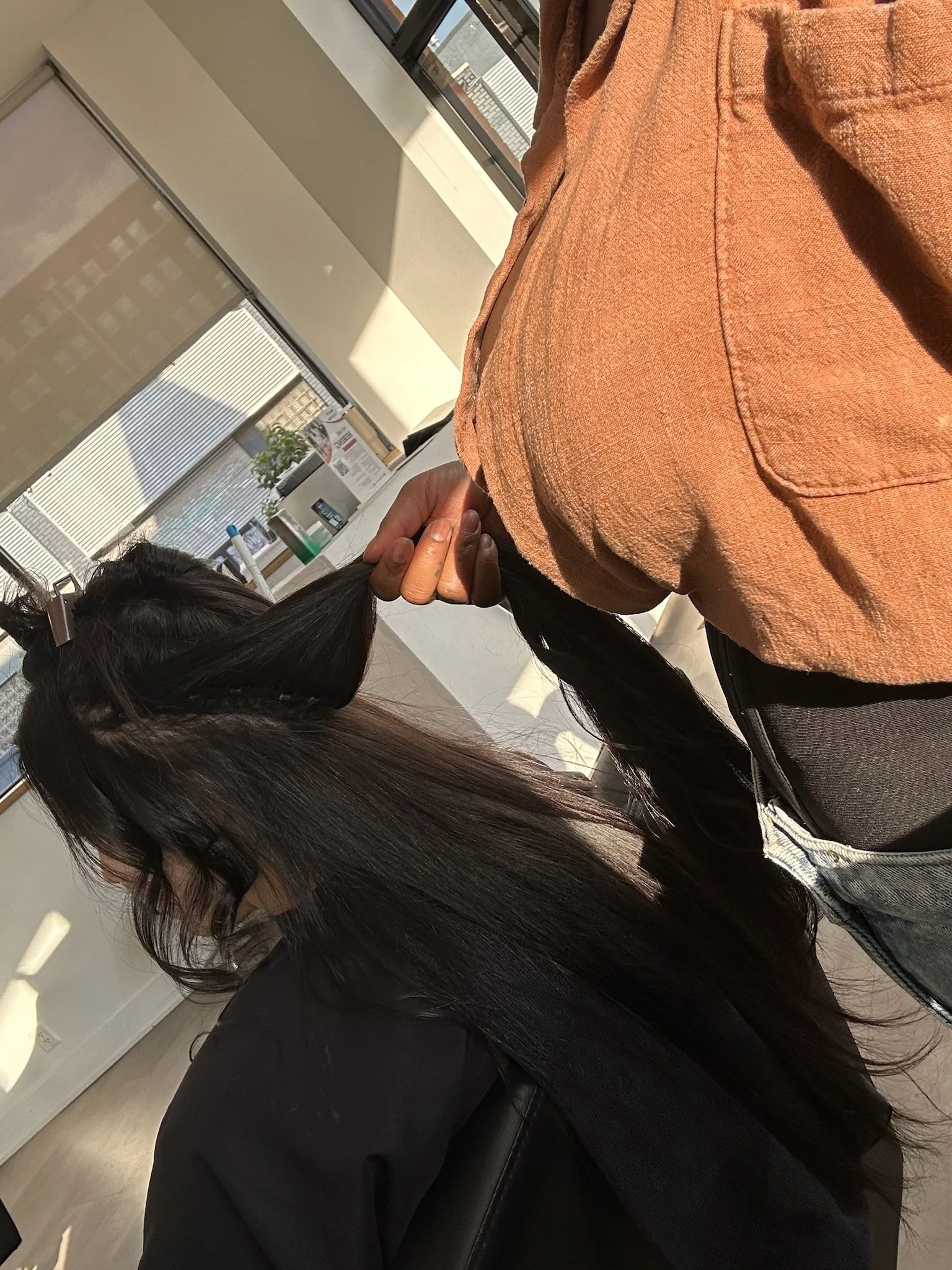 A stylist in an orange shirt holds a section of long dark hair above a black salon cape.