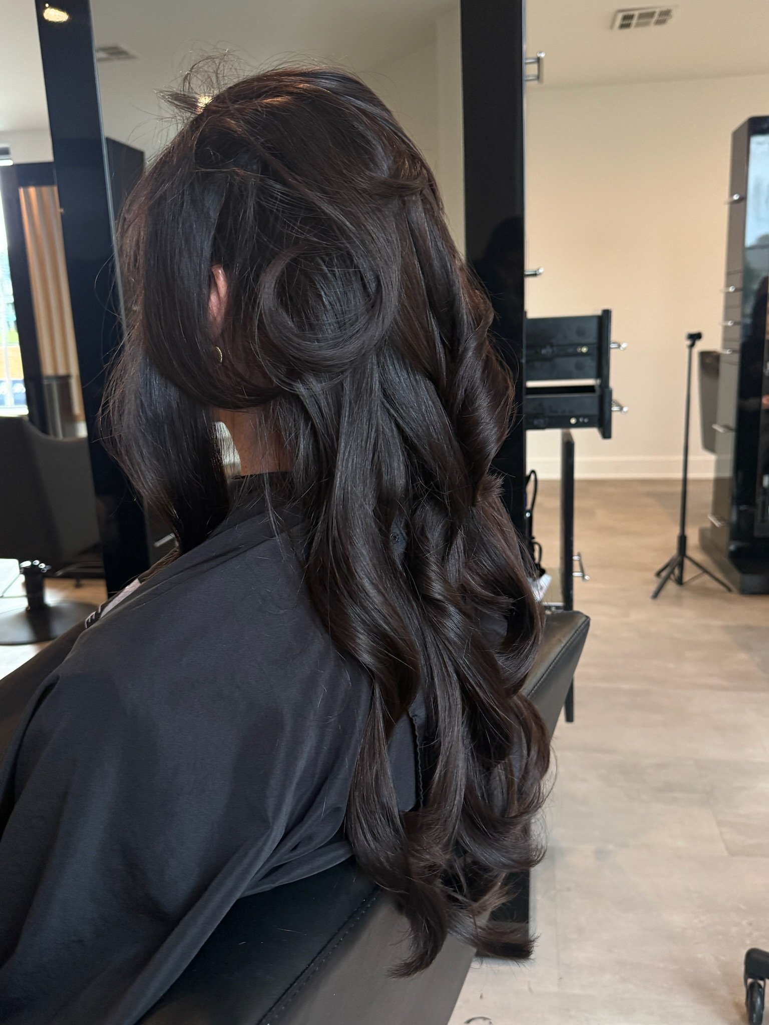 Long dark hair arranged in large curls over a black salon cape, viewed from the side and back.