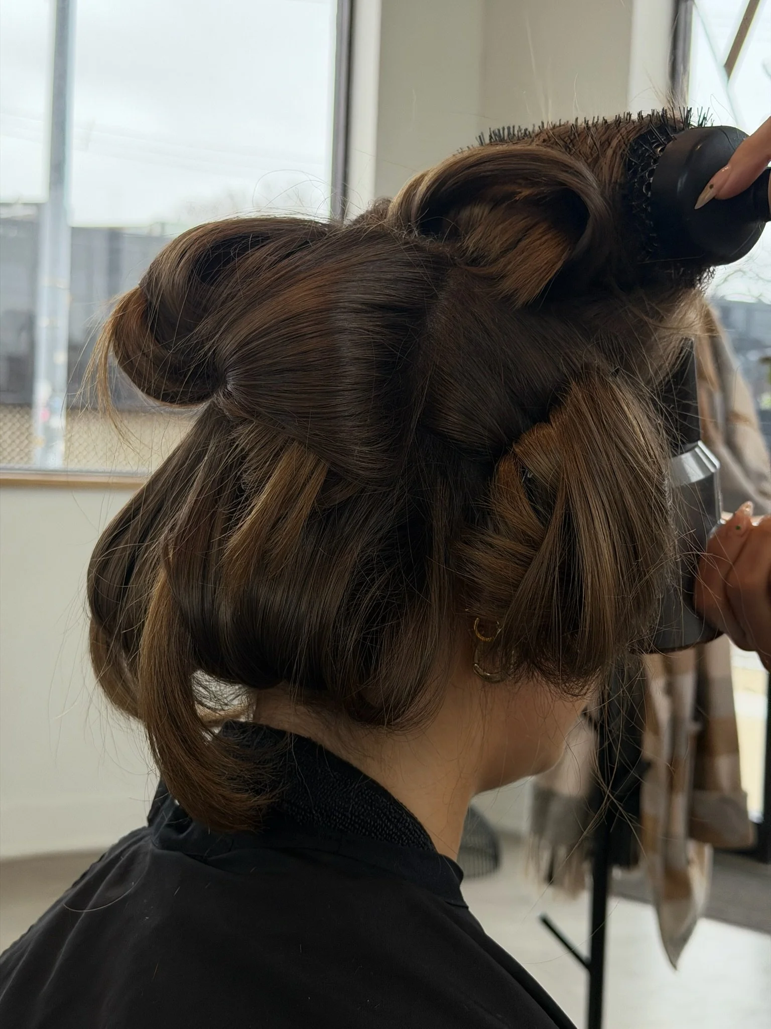 Sectioned brown hair being shaped with a round brush near a salon window.